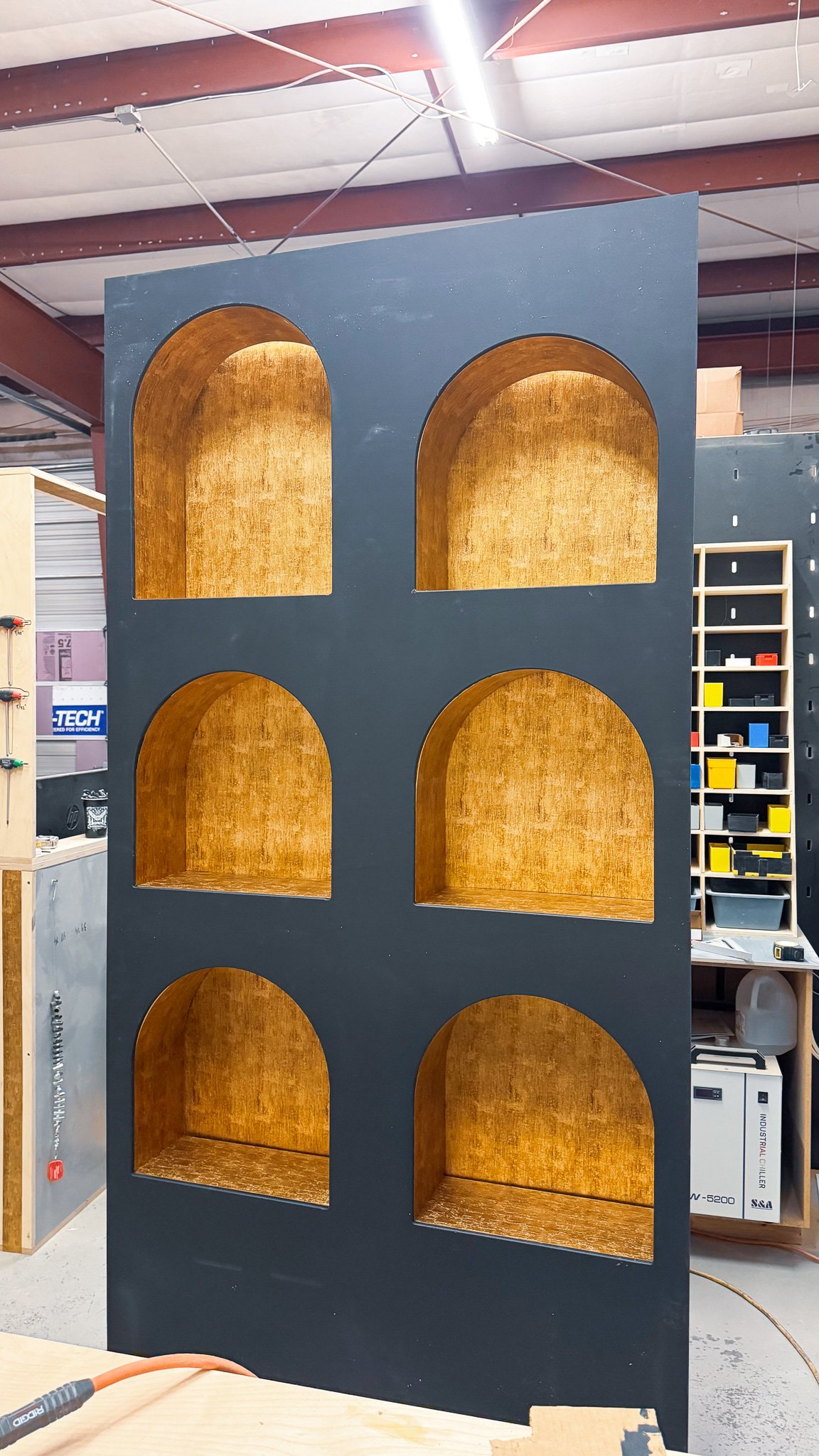 A black multi-level wooden bookshelf with arched openings on each shelf, inside is a gold-colored finish. The background shows a workshop or storage area with shelves, tools, and machinery.