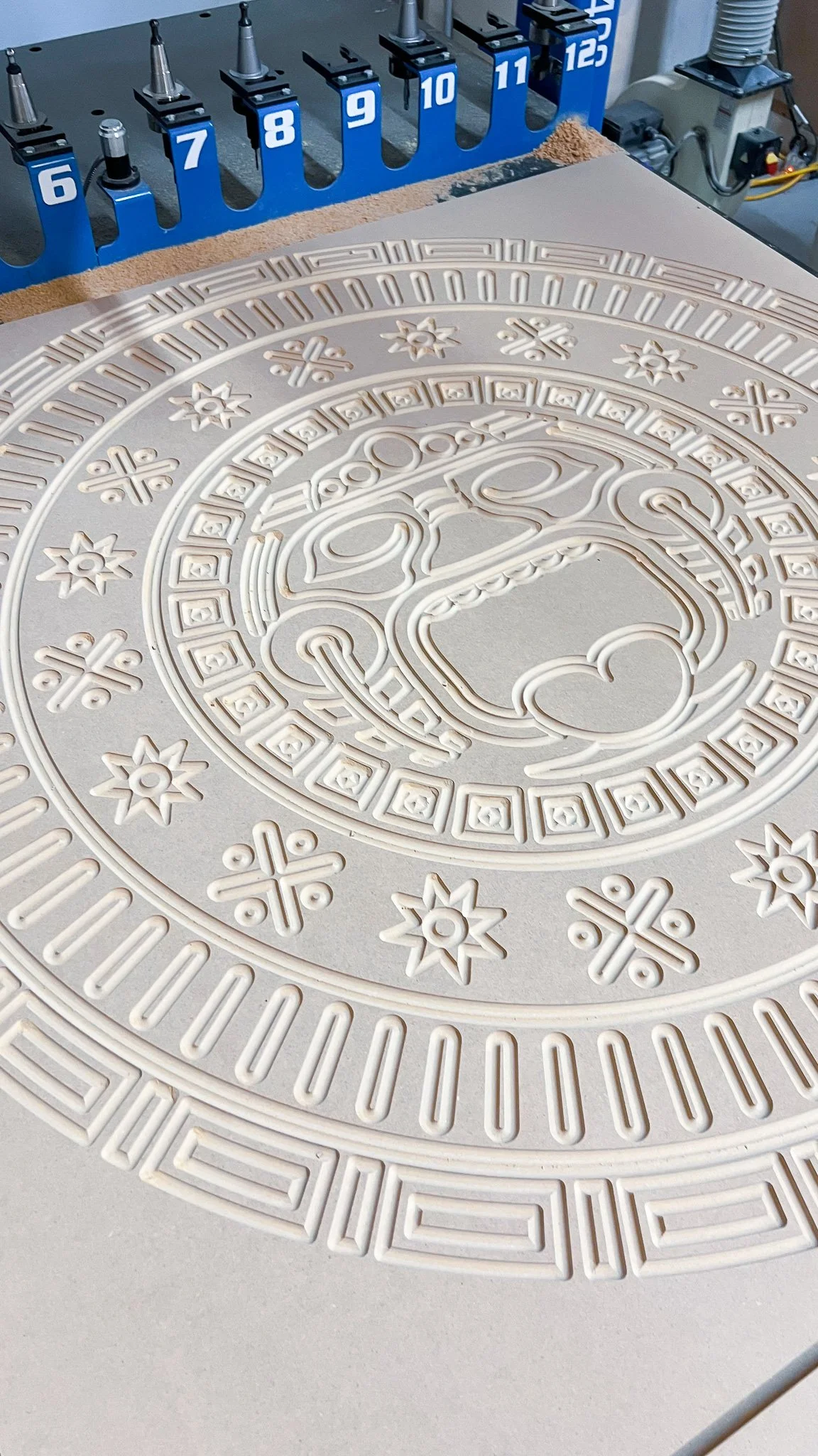 Close-up of a carved circular design on a white surface, with a woodworking setup in the background including clamps numbered 6 to 12 and some machinery.
