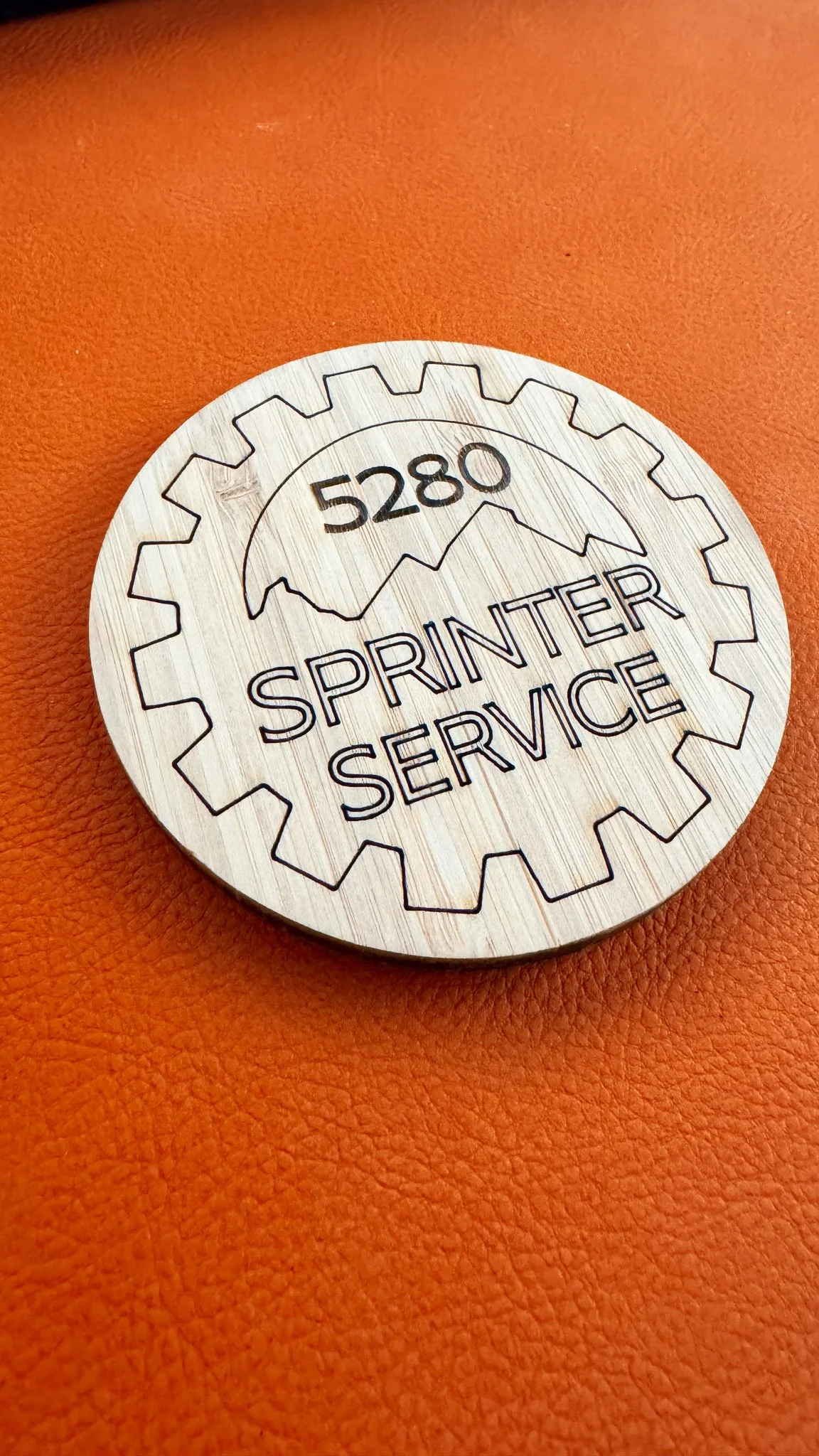 Circular metal license plate or badge with a gear contour, engraved with the number 5280, mountain graphic, and the words 'SPRINTER SERVICE' on an orange textured surface.