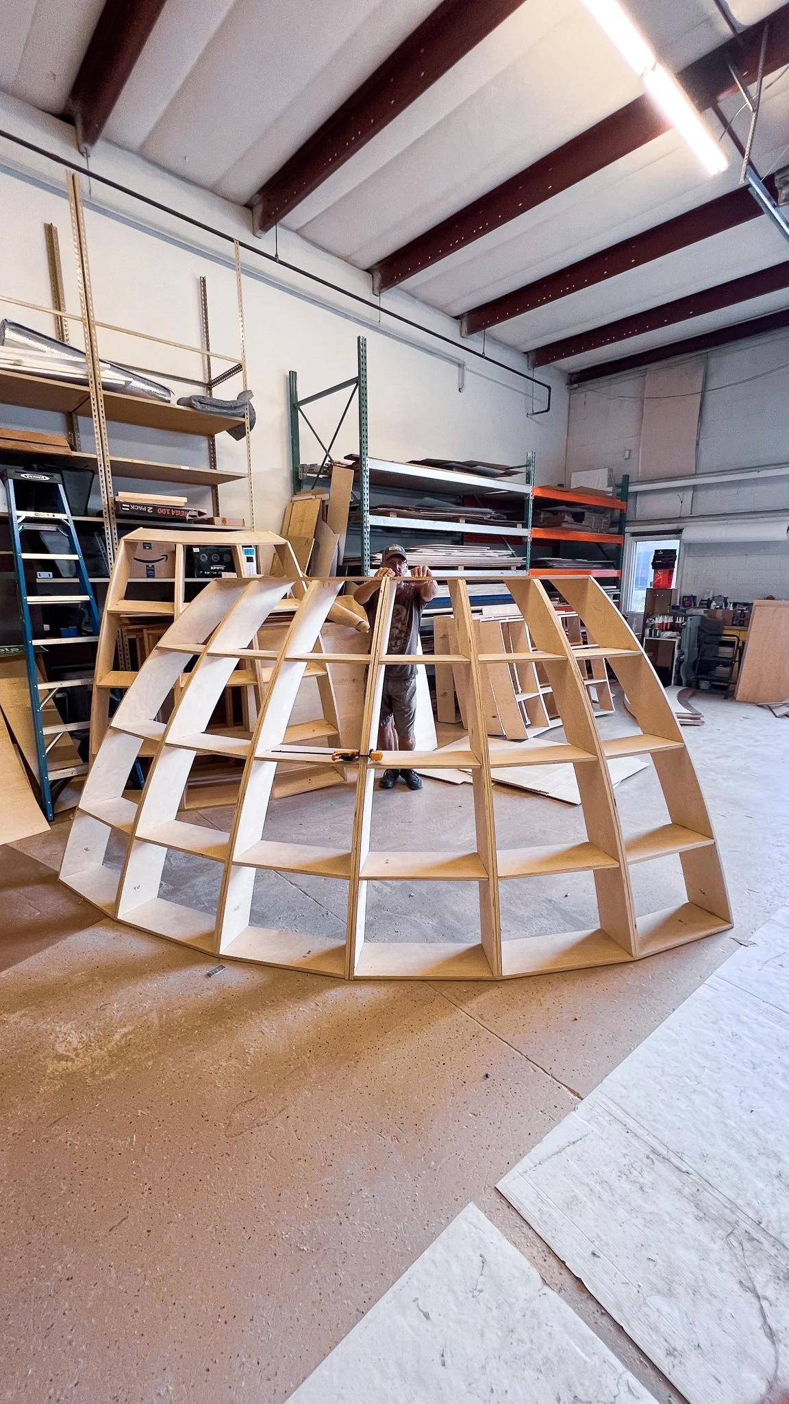 A person working in a woodworking shop with scaffolding and wooden shelves, assembling a large, curved wooden structure.