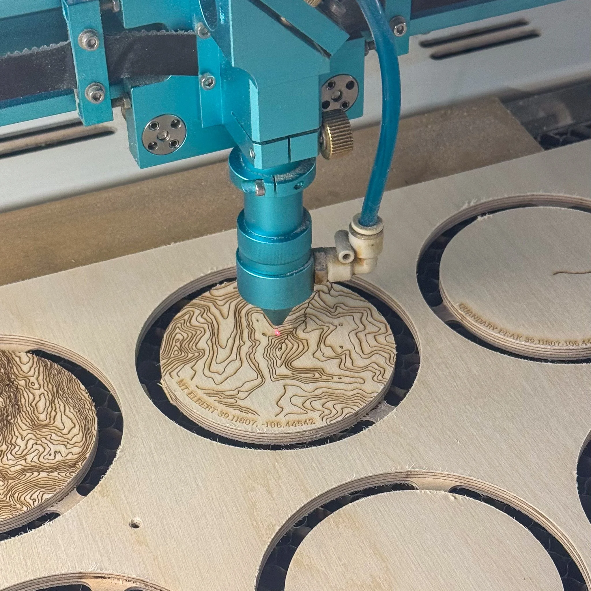 A laser engraver is carving a topographic map into a circular piece of wood. The engraving machine is blue and positioned above the wood, with a red laser beam focused on the surface.