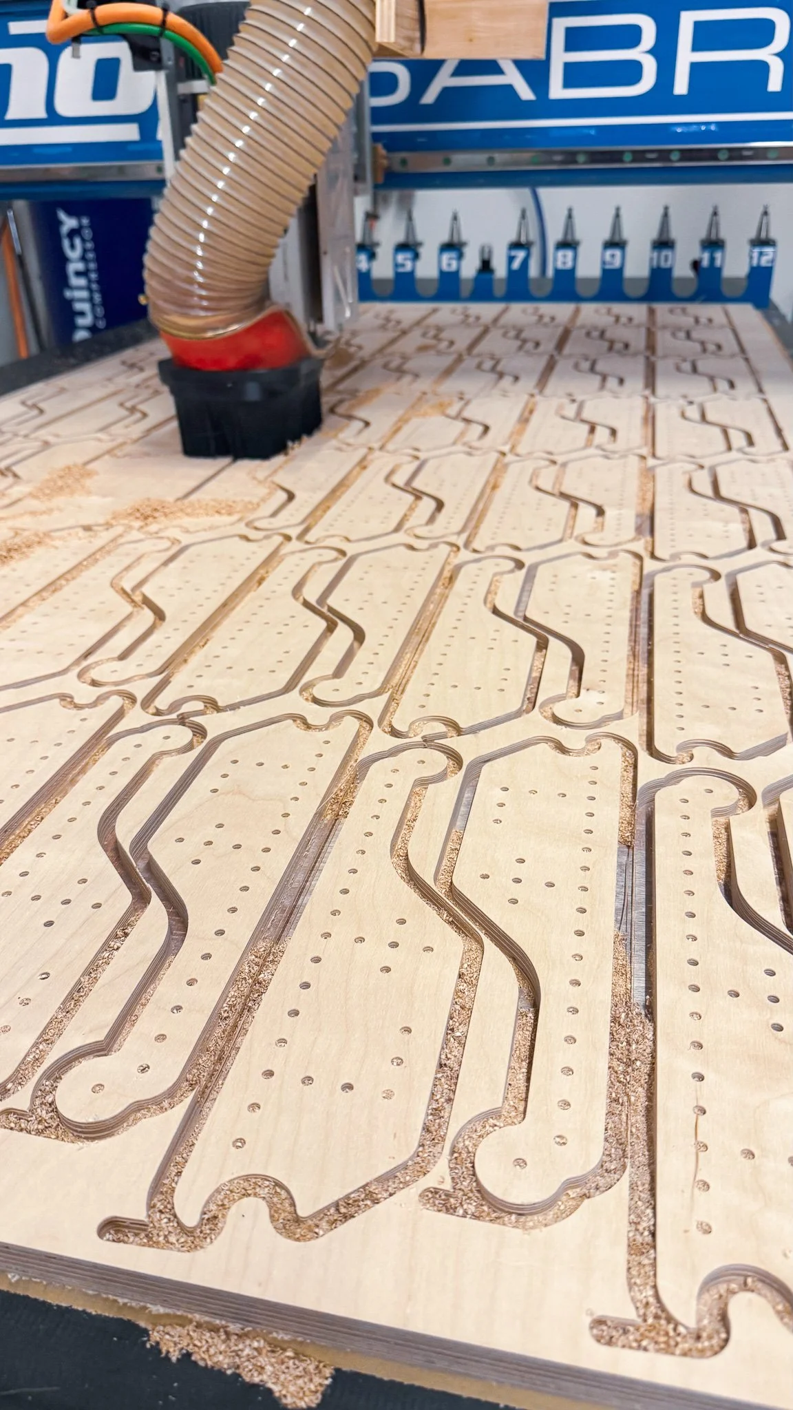 Laser cutting machine creating a maze pattern on a sheet of wood. The machine has a flexible hose and numbered stations in the background.