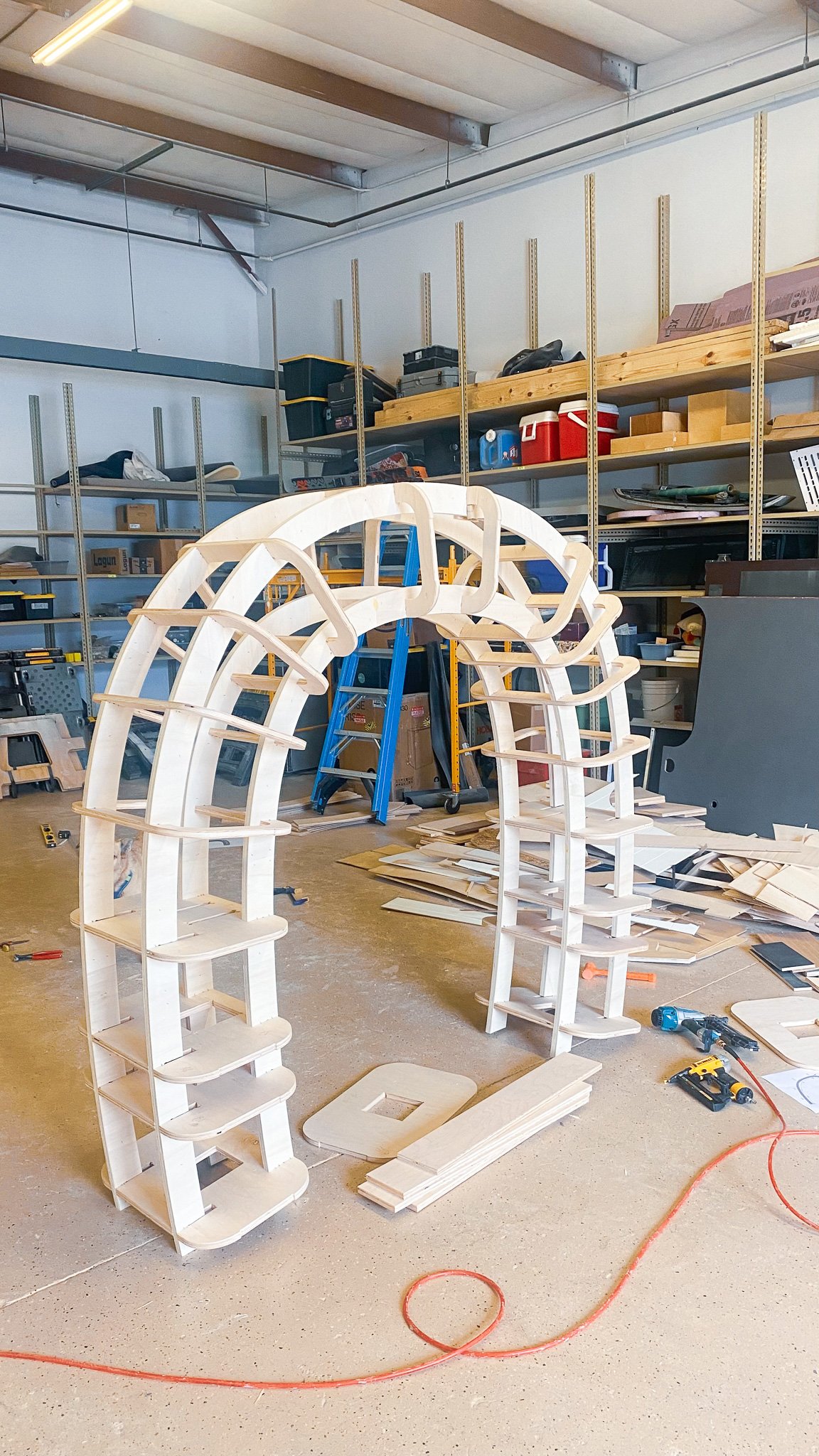 A workshop with wooden arch structures in progress, tools, and materials scattered around. Metal shelving with various supplies in the background.