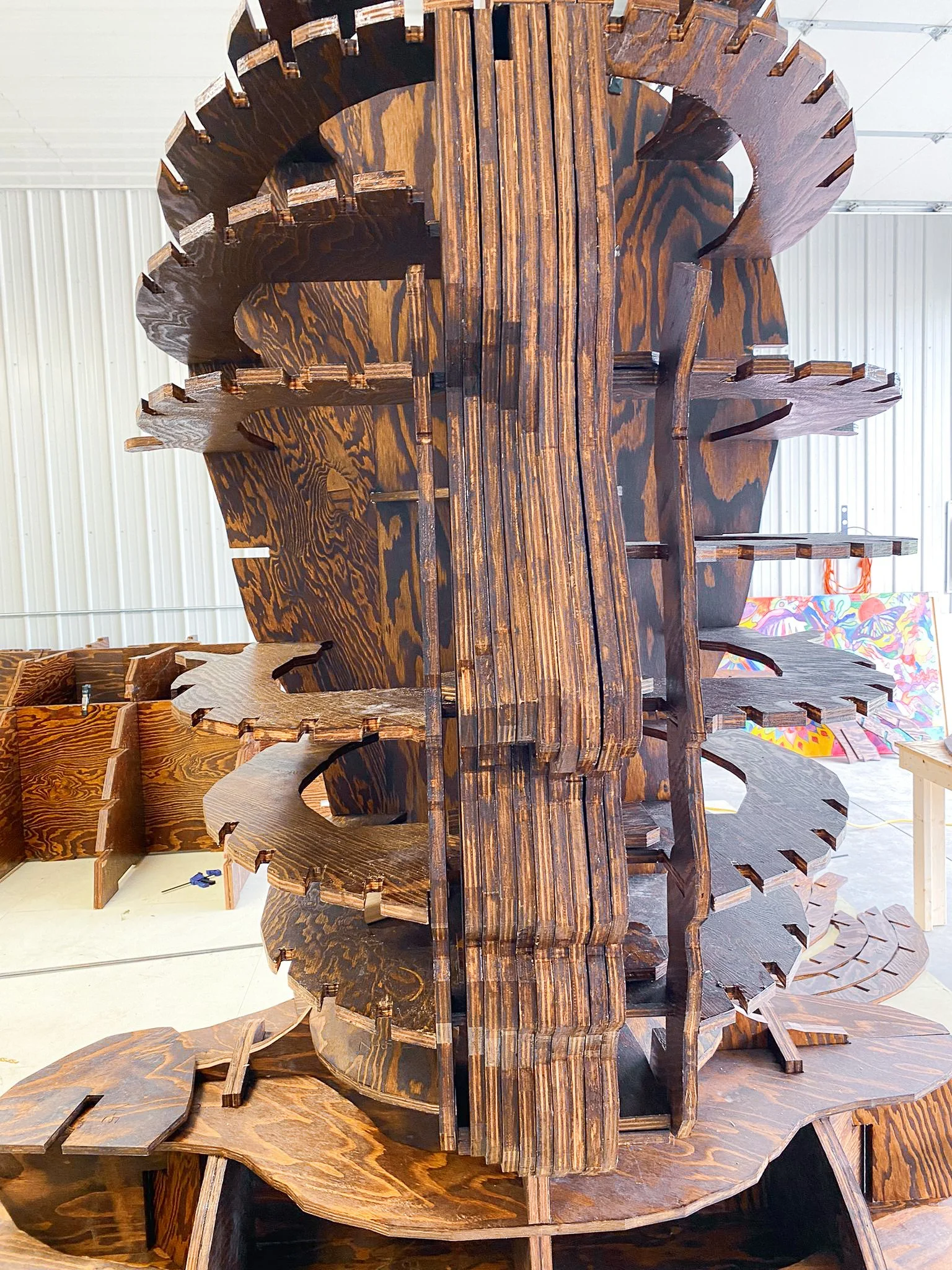 A large wooden structure resembling a gear or cog with intricate cutouts and a polished surface, displayed indoors with a white ceiling and colorful background.