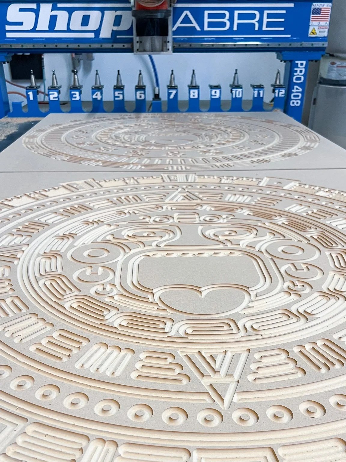 Two intricate, circular, carved patterns on flat surfaces on top of a Shop Sabre CNC router machine.