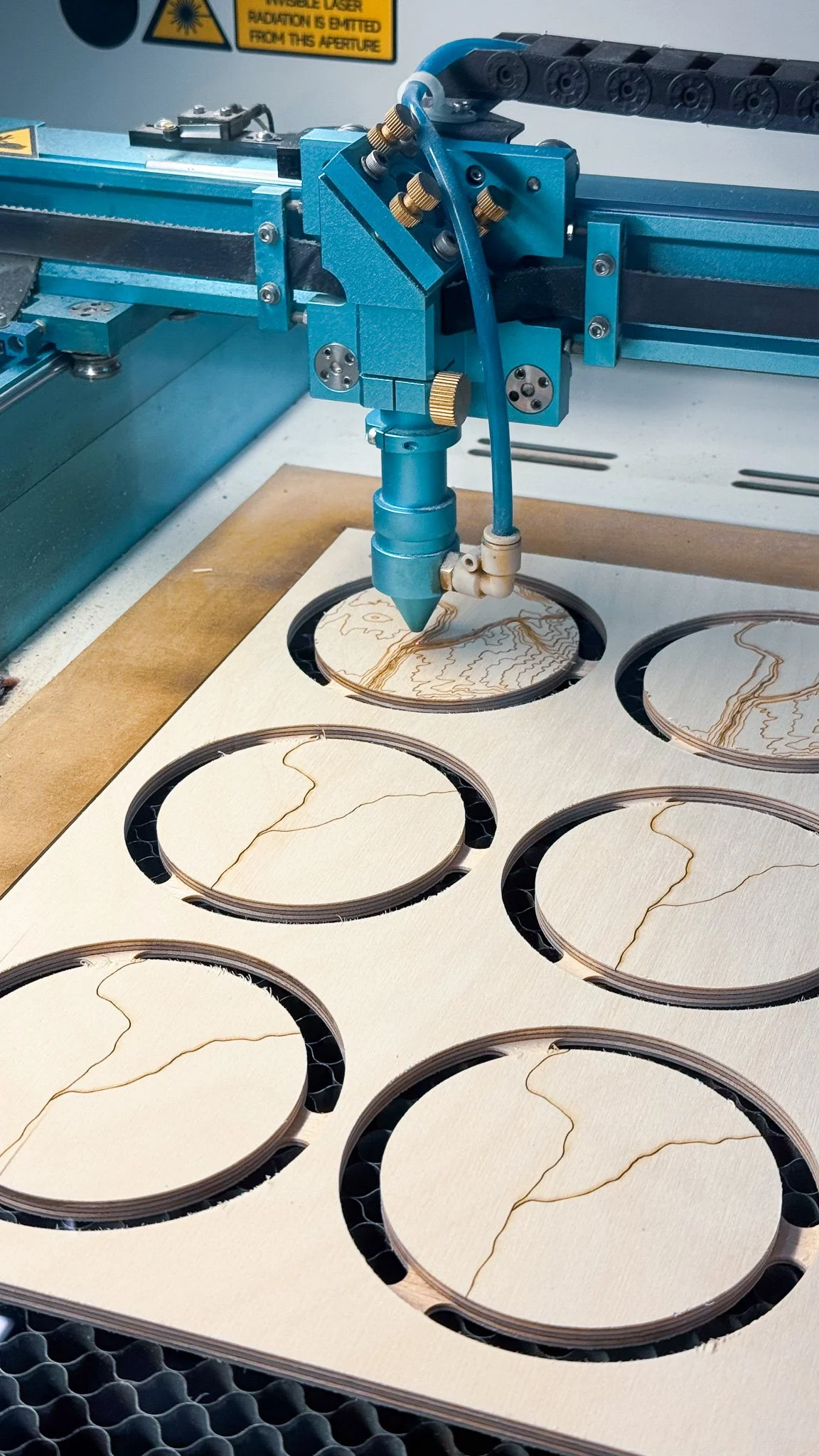 A laser cutter machine engraving cracks and patterns on wooden circles placed on a perforated surface.