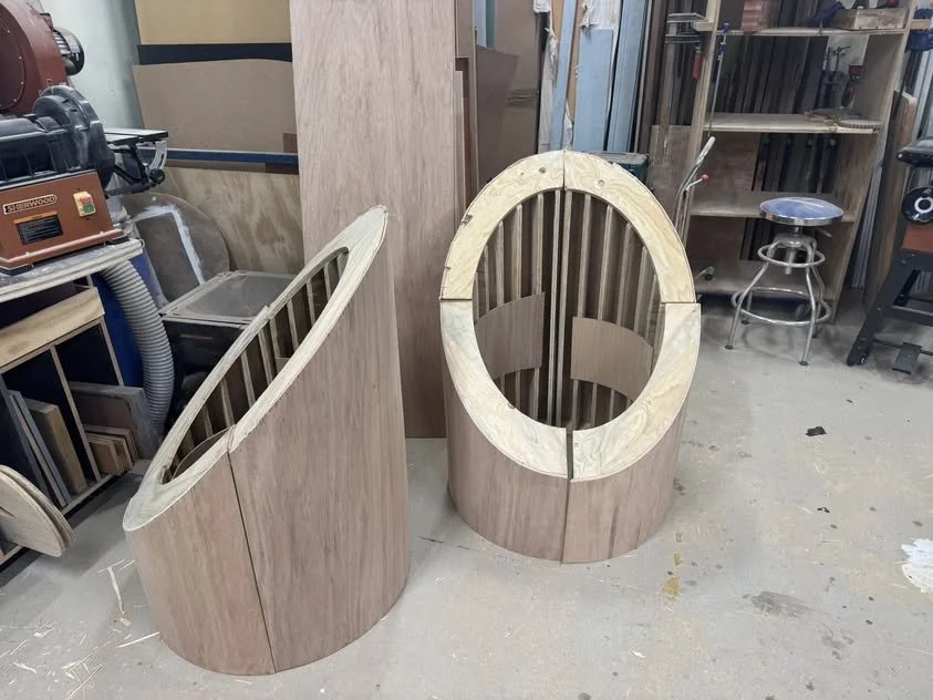 Two wooden oval-shaped furniture pieces in a woodworking shop, with one piece showing an open top revealing internal wooden slats.