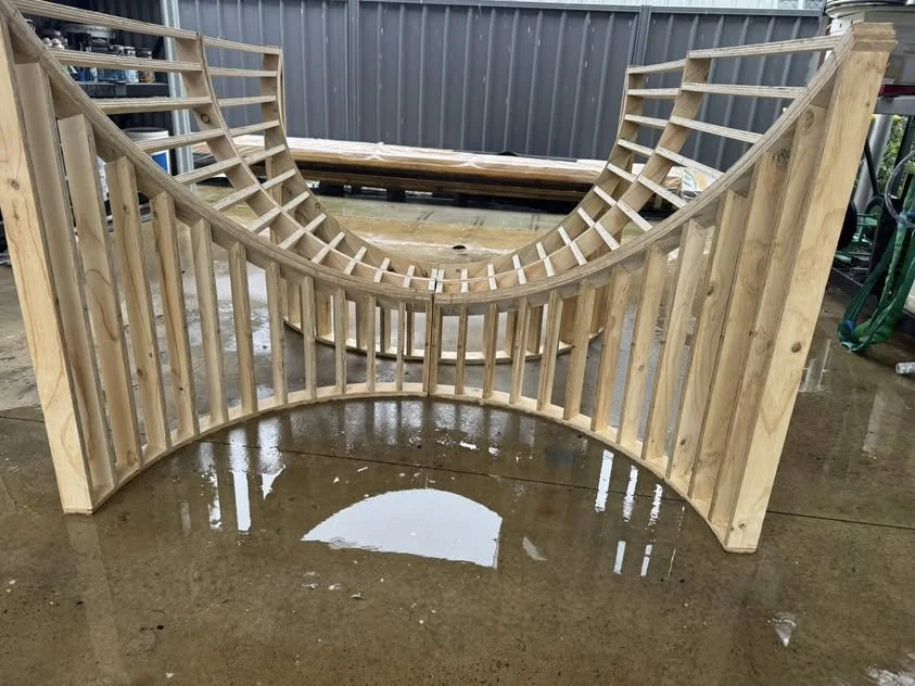 A wooden skate ramp in progress with curved sides and a semi-circular shape, placed on a wet concrete ground outdoors.