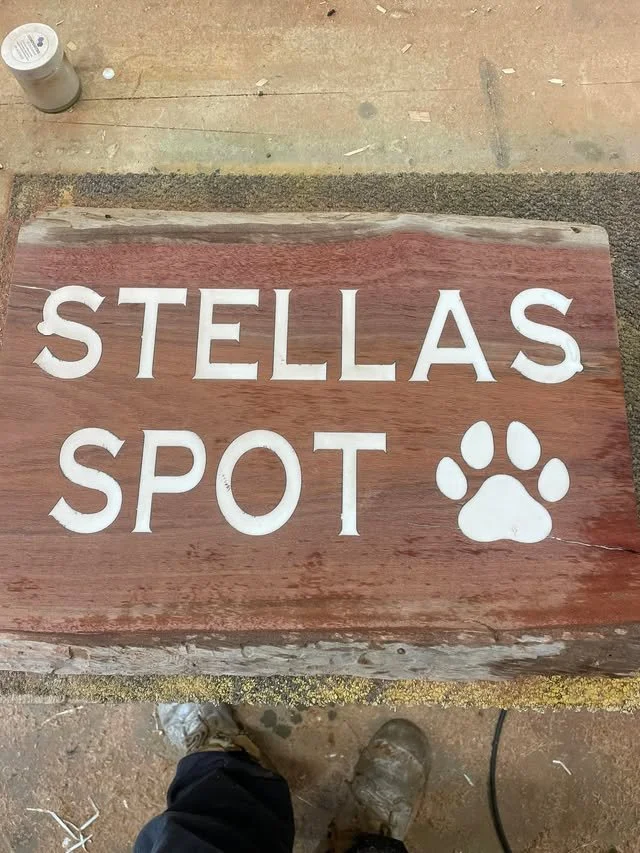 A wooden sign with white letters that reads 'STELLAS SPOT' and a paw print symbol, placed on a wooden surface with a person standing nearby wearing boots.
