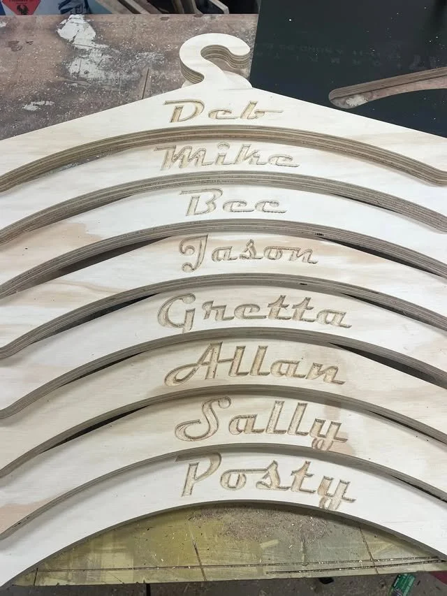 A stack of wooden signs with curved tops, each featuring engraved names of popular American sitcom characters, including DArlt, Mike, Bee, Jasun, Ghettra, Allain, Sallur, and Postur.