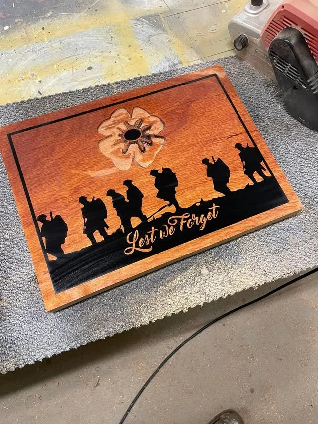 Wooden plaque with cutout silhouettes of soldiers walking, a flower shape at the top, and the words "Lest we Forget" engraved at the bottom.