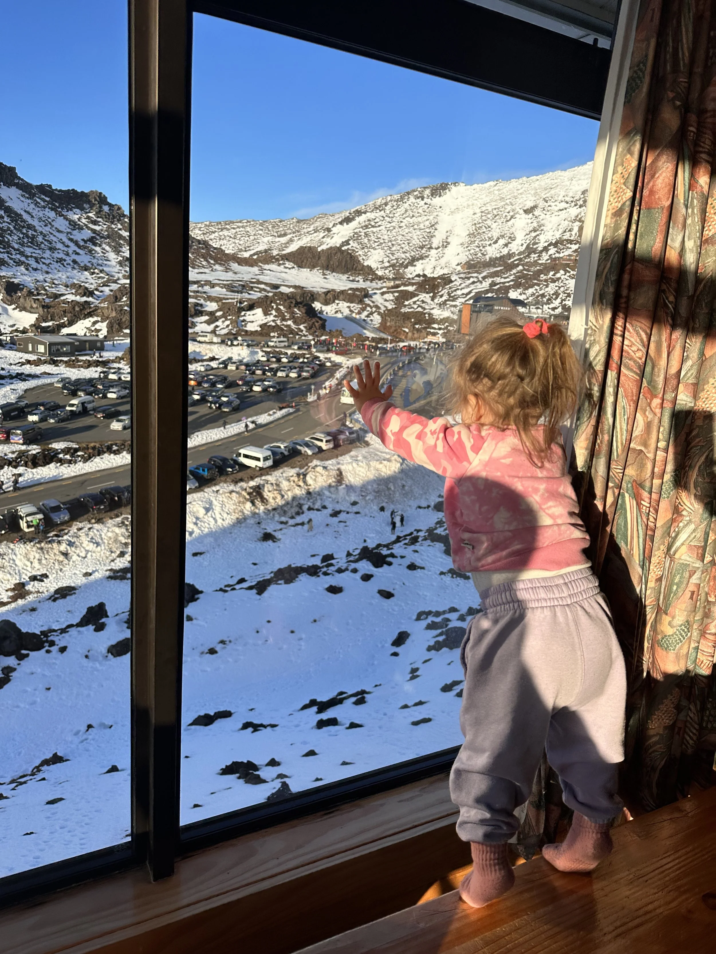 A young girl with curly hair in a pink hoodie and gray sweatpants standing barefoot on a wooden floor, touching a window while looking outside at snow-covered mountains and a parking lot below on a clear, sunny day.