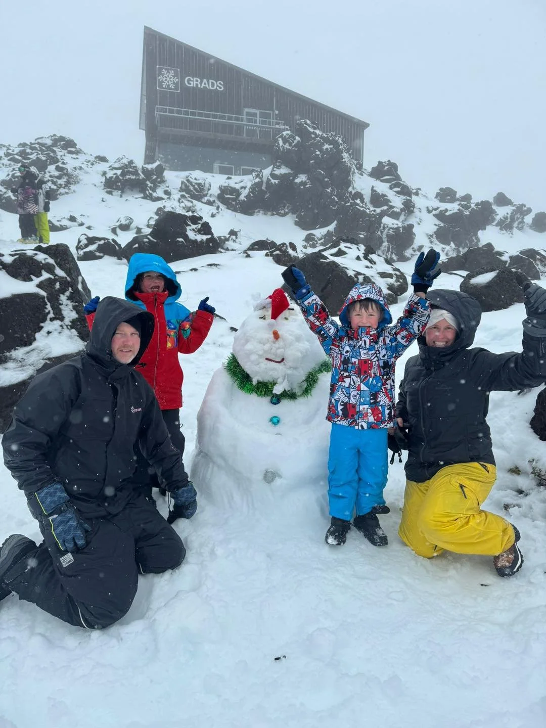 Group of four children and adults outside in snow, with a snowman decorated with a red hat, green tinsel, and a face. They are dressed in winter clothing, raising their hands and smiling, with a rocky, snowy landscape and a building labeled 'Grads' in the background.