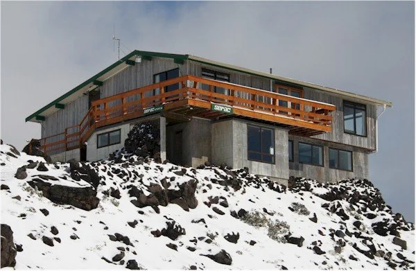 A wooden house on a snow-covered rocky hillside, with large windows and a balcony.