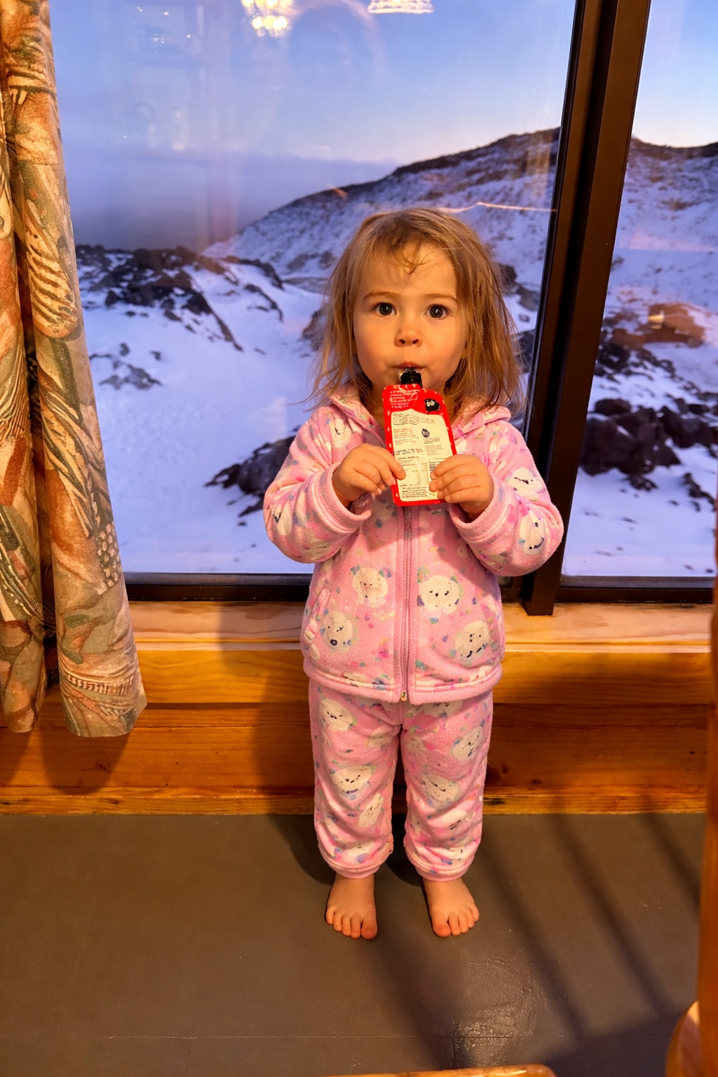 A young girl in pink pajamas with a fleece jacket and pants featuring a snowman pattern, holding a juice box with a straw, standing by a window with a snowy mountain landscape in the background.
