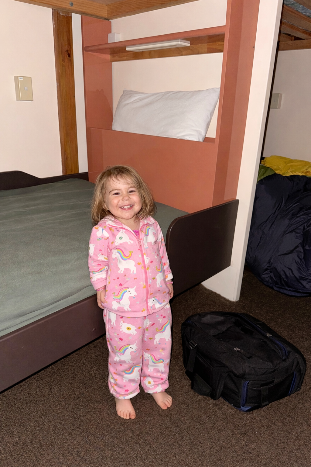 A young girl standing in pajamas with unicorn and rainbow print, smiling in front of a bed with a pink headboard and a bedroom wall, with a black backpack on the brown carpeted floor.