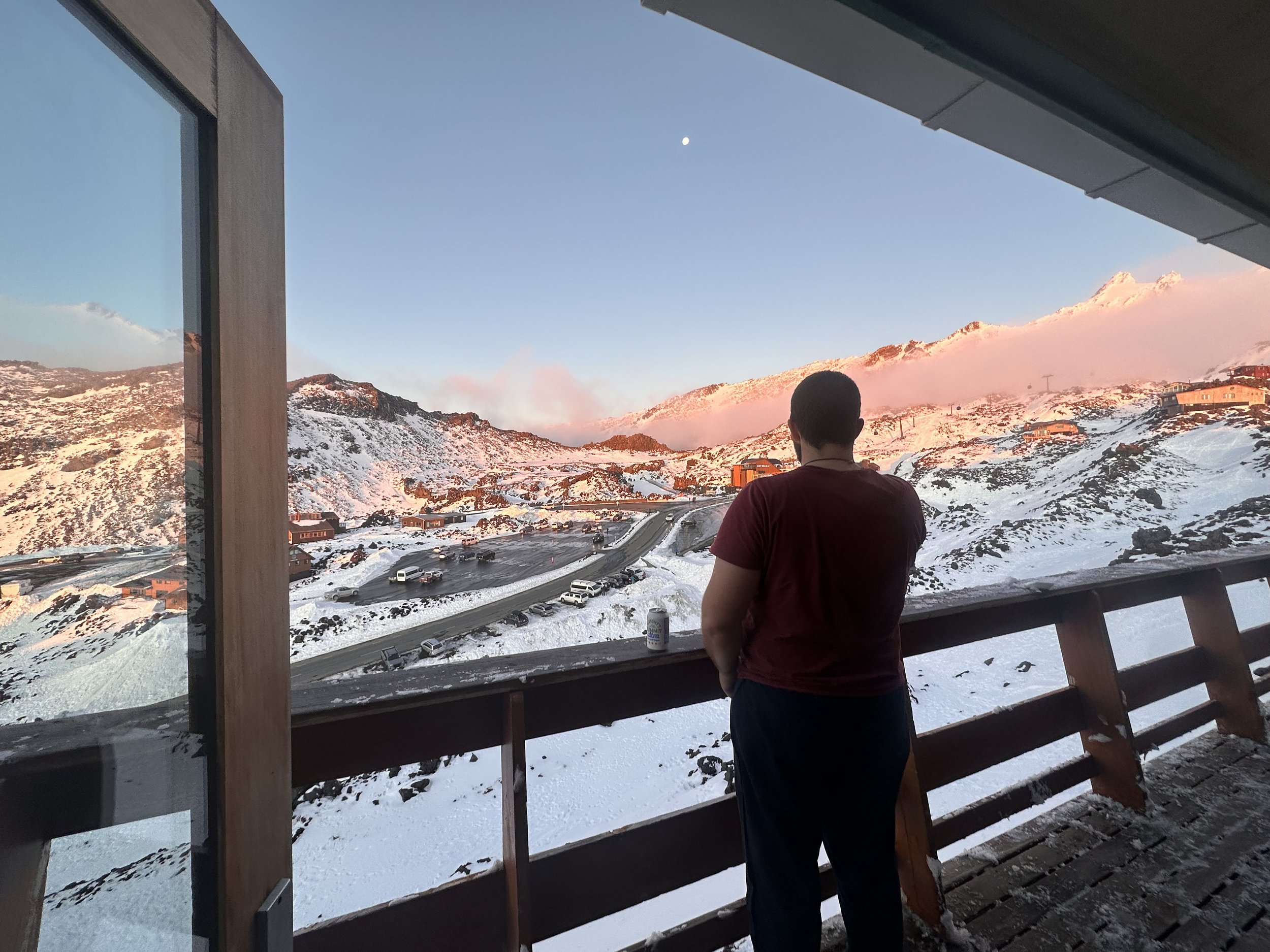 A person with short dark hair wearing a red shirt is standing on a balcony with a snow-covered mountain landscape in the background during sunset, with the moon visible in the sky.