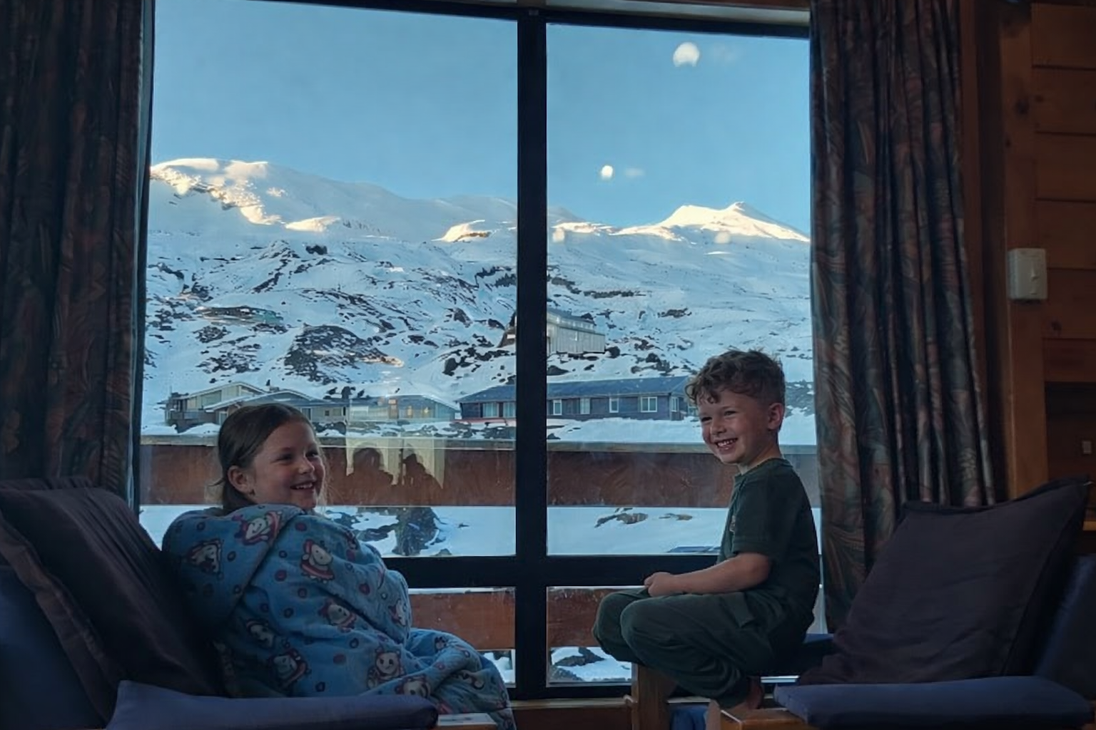 Two children smiling and sitting inside a room near a large window with a snowy mountain landscape outside.