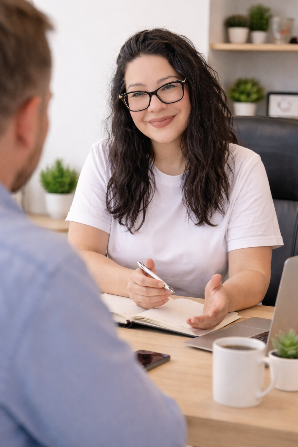 A woman with glasses and wavy dark hair smiling and talking to a man in an office or meeting room, with a notebook, pen, laptop, coffee mug, and plants visible.