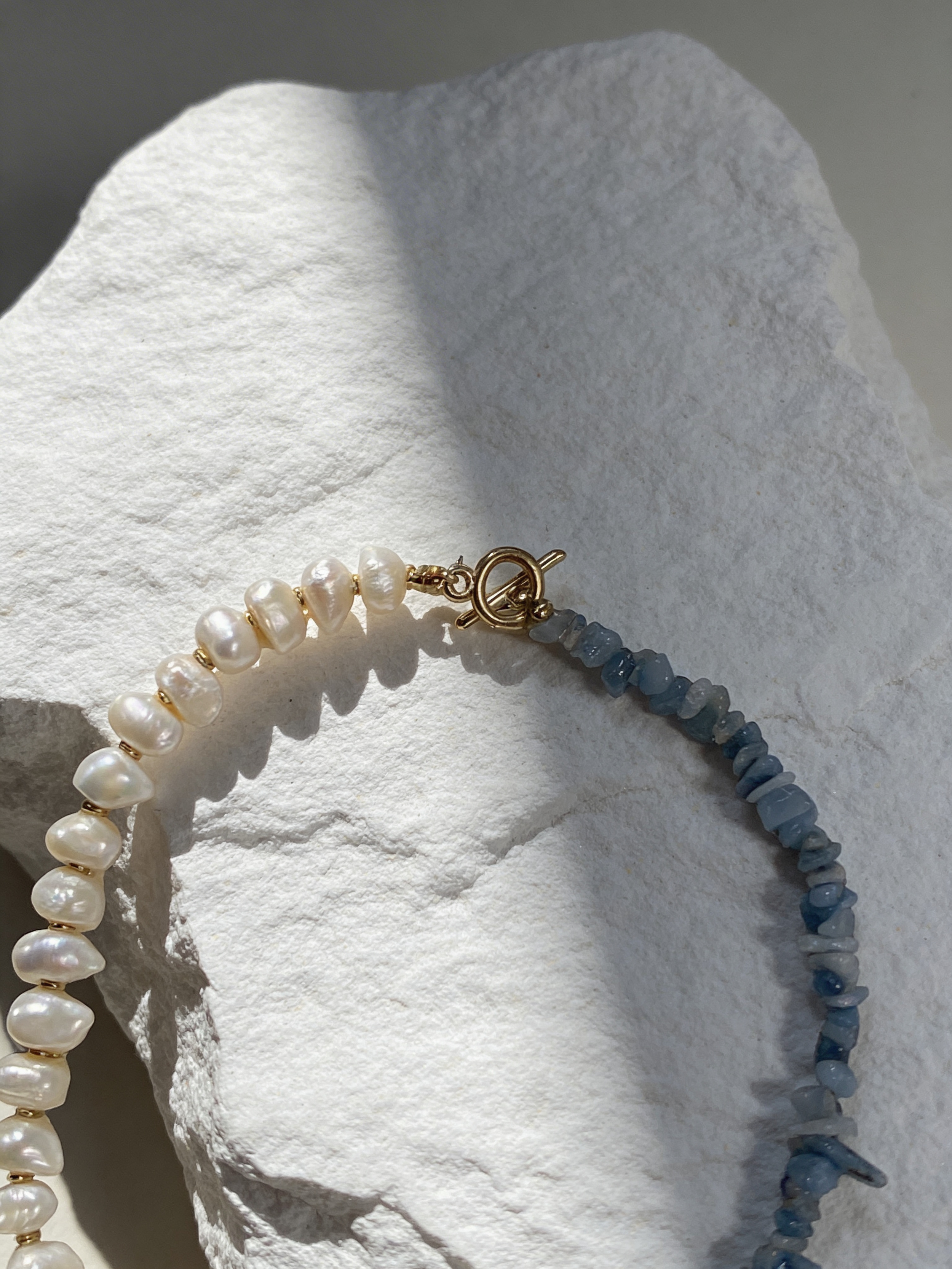 Close-up of a pearl and blue stone beaded bracelet with a gold toggle clasp resting on a textured white stone.