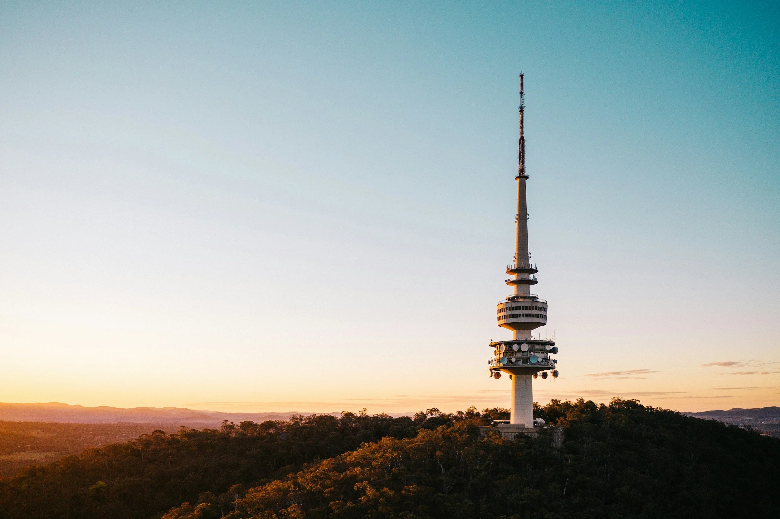A tall communication tower on a hill during sunset, with a clear sky above and trees covering the hill.