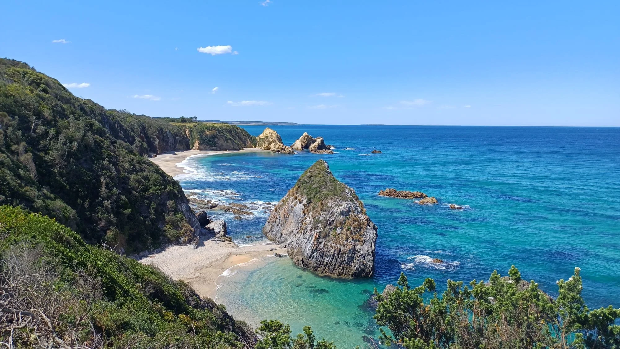 A scenic coastal view with a sandy beach, large rocks in the water, and lush green cliffs under a bright blue sky.