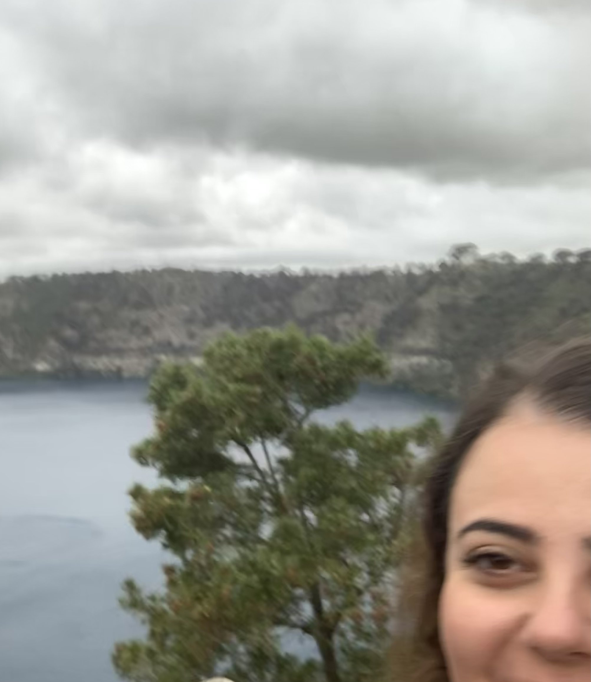 Partially visible smiling woman in front of a scenic outdoor landscape with a body of water, trees, hillside, and cloudy sky.