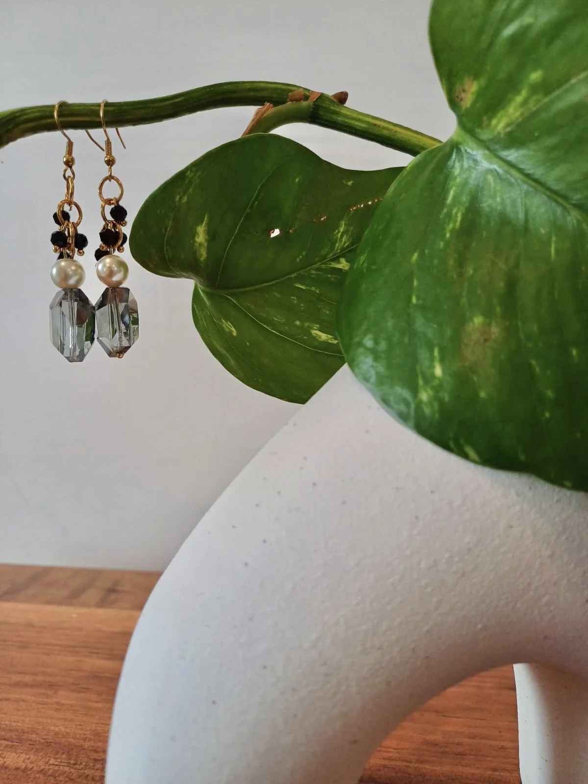 A white ceramic vase with a large green leaf, small holes, and yellow variegation. A pair of black, gold, pearl, and crystal dangle earrings hanging from a green stem with leaves in the background, on a wooden surface.