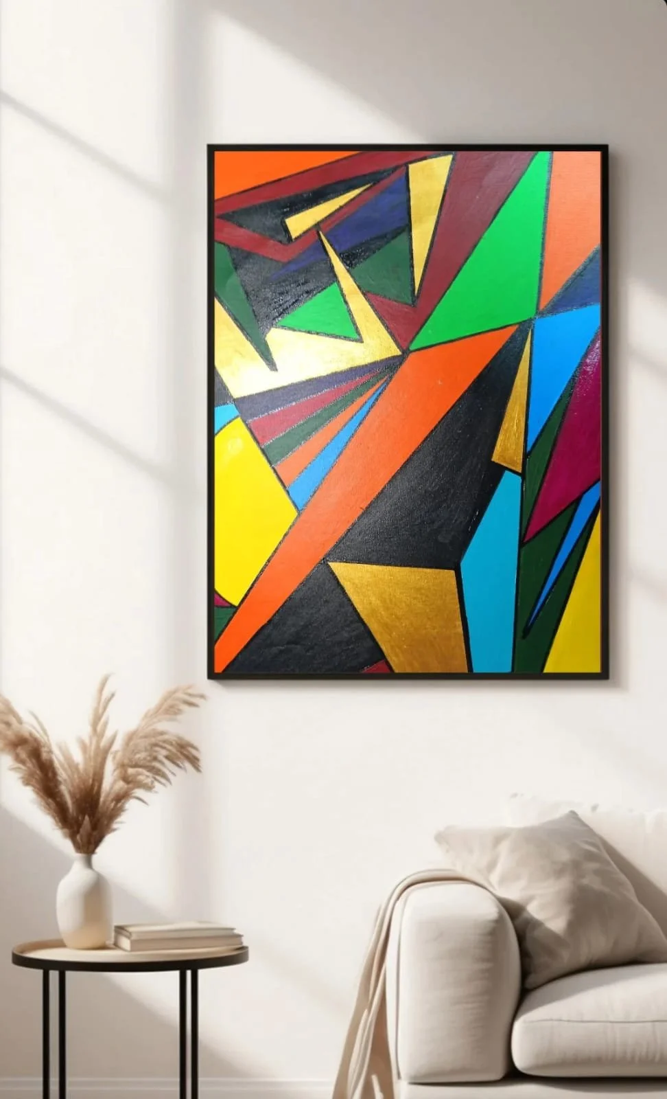 Un dipinto astratto con forme geometriche colorate appeso a una parete bianca, parte di un soggiorno con un divano beige, un tavolino con una pianta di pampas e alcune riviste.