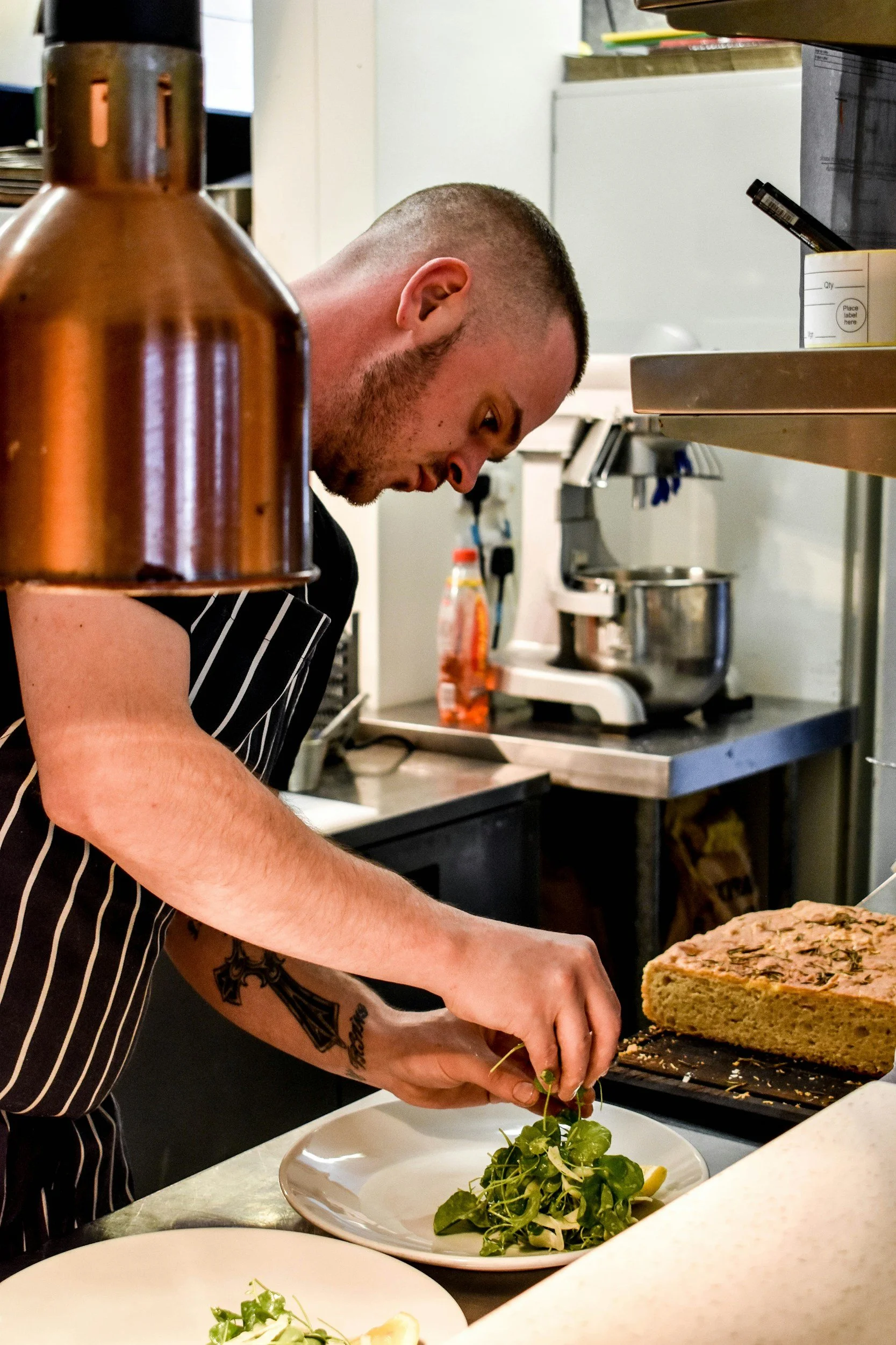 A man with a beard and tattoos wearing a striped apron is preparing food in a kitchen. He is leaning over a counter, garnishing a salad with microgreens and lemon slices on a white plate. A large cake with a textured topping is also on the counter. In the background, there are kitchen appliances, shelves, and utensils.