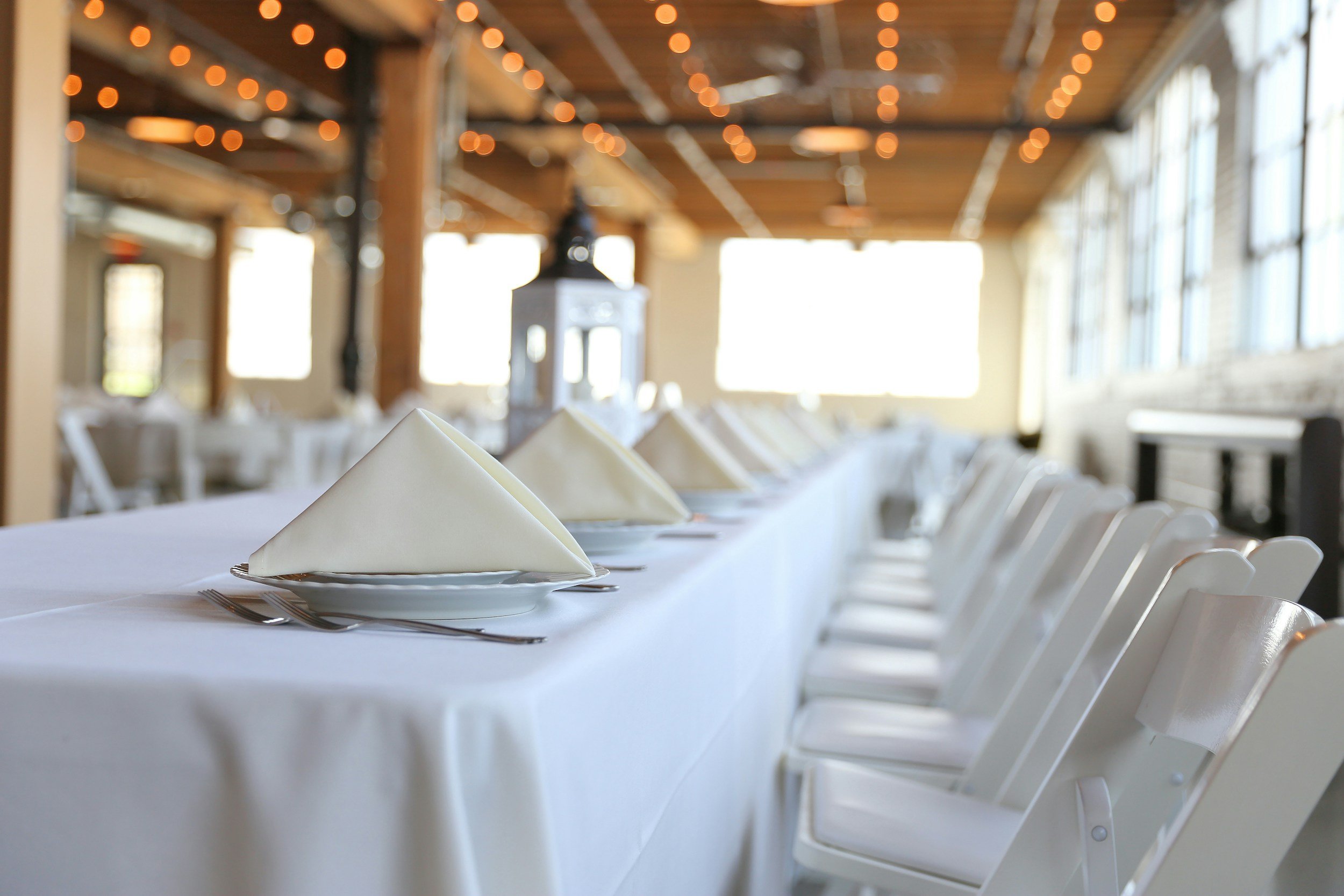 Banquet table with white tablecloths, set with white napkins folded into triangles, silverware, and empty white plates, in a well-lit room with large windows, wooden beams, and string lights on the ceiling.