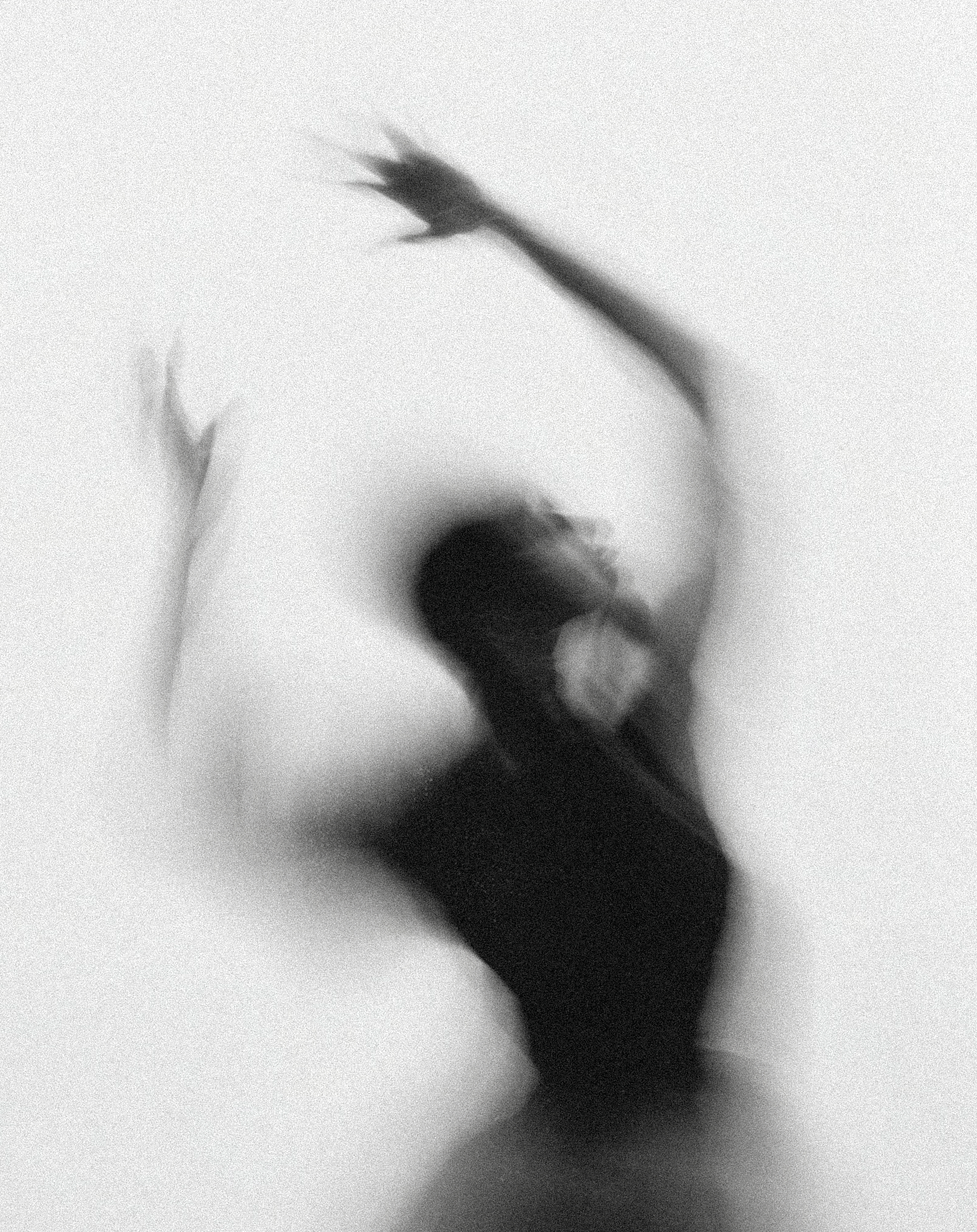 A blurred black and white image of a person dancing with arms raised, standing against a plain background.