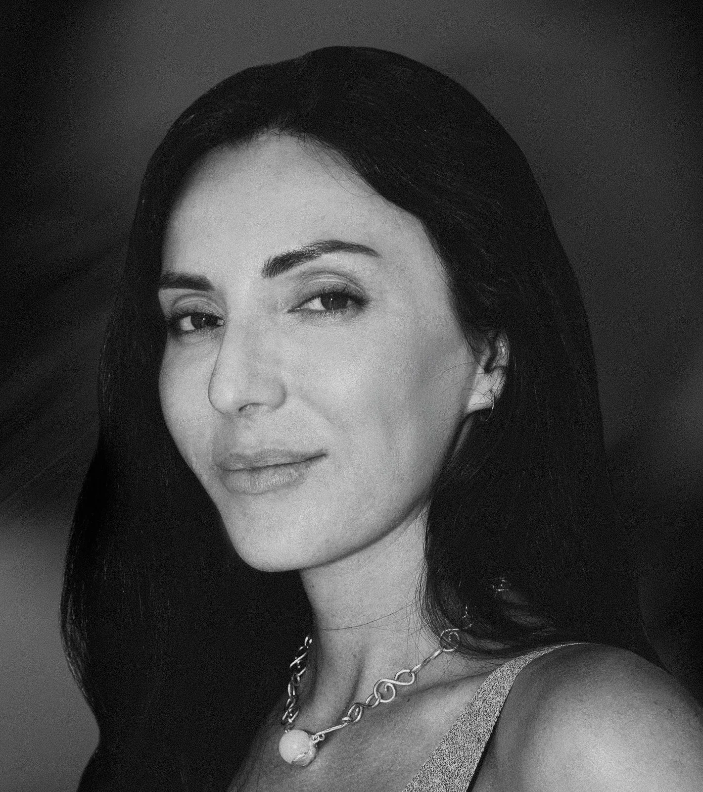 A black and white portrait of a woman with long dark hair, wearing a necklace and a sleeveless top, looking at the camera with a slight smile.