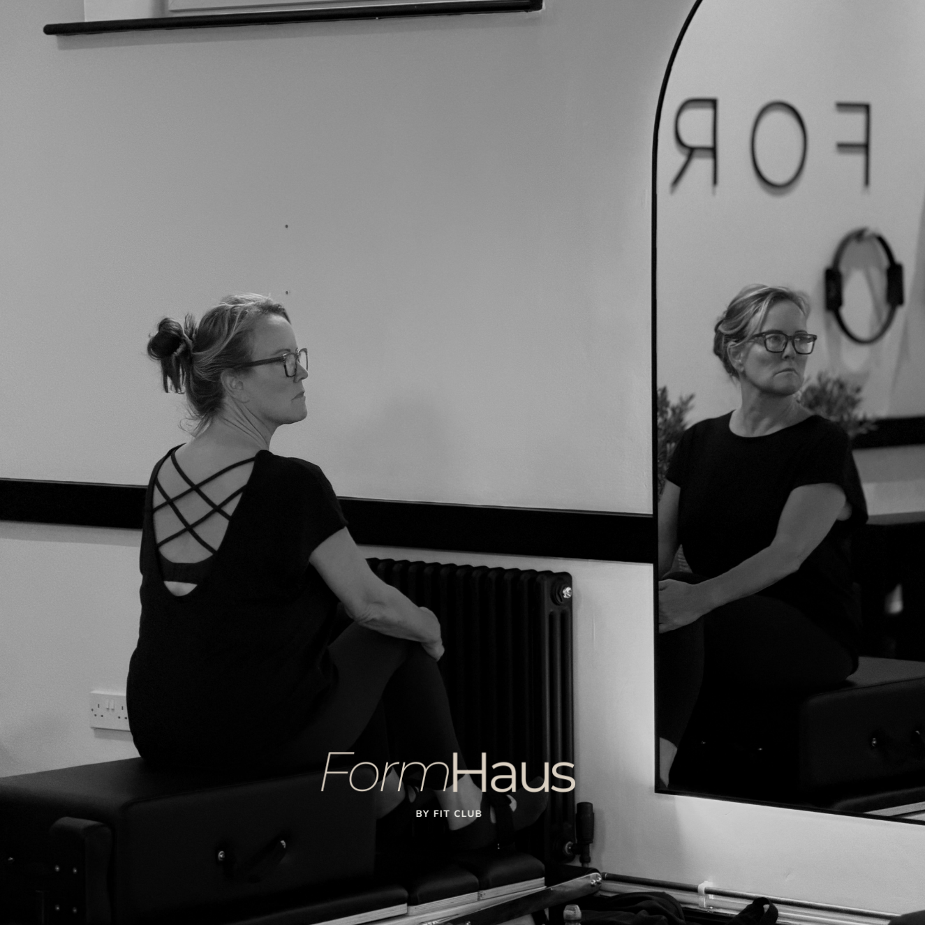 A woman with glasses and a black top with crisscross straps on the back, sitting on a workout bench, looking at her reflection in a mirror in a fitness studio.