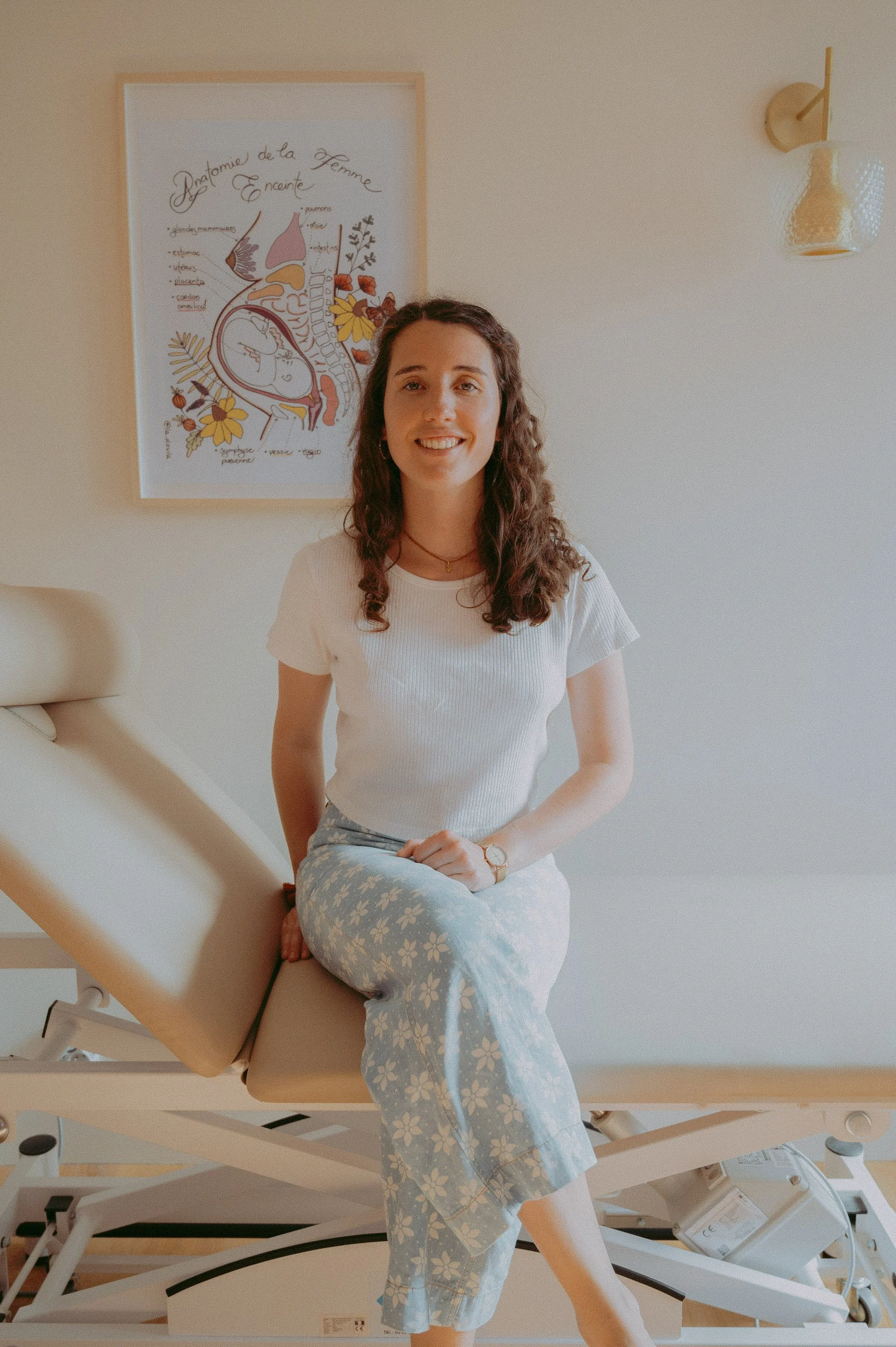 Une femme souriante assise sur une table d'examen médical, portant un t-shirt blanc et un pantalon à motifs floraux, dans une pièce lumineuse avec un poster médical sur le mur et un éclairage mural.