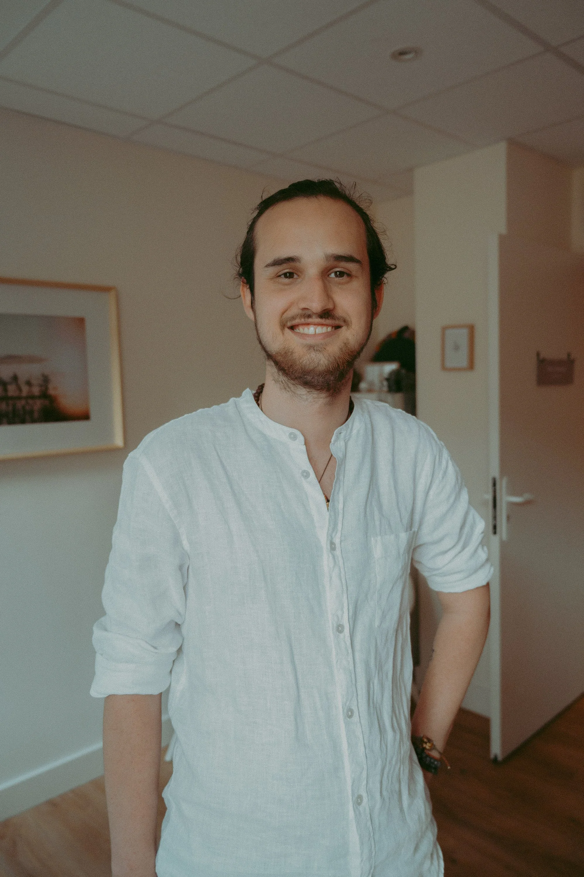 Un homme souriant avec une barbe légère, portant une chemise blanche avec les manches retroussées, debout dans une pièce intérieure