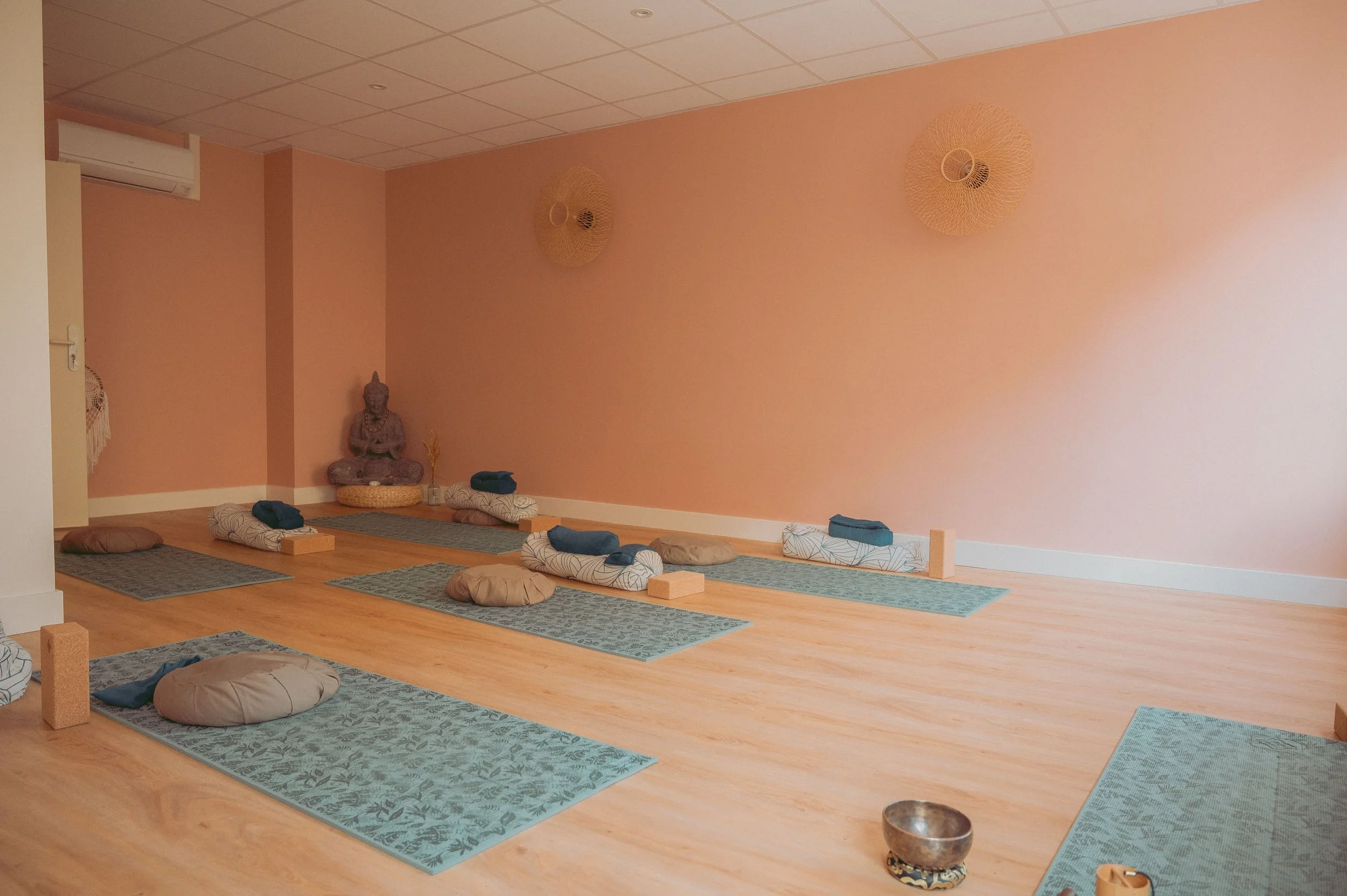 Salle de yoga avec des tapis, des coussins et une statue de Bouddha contre un mur rose pastel.