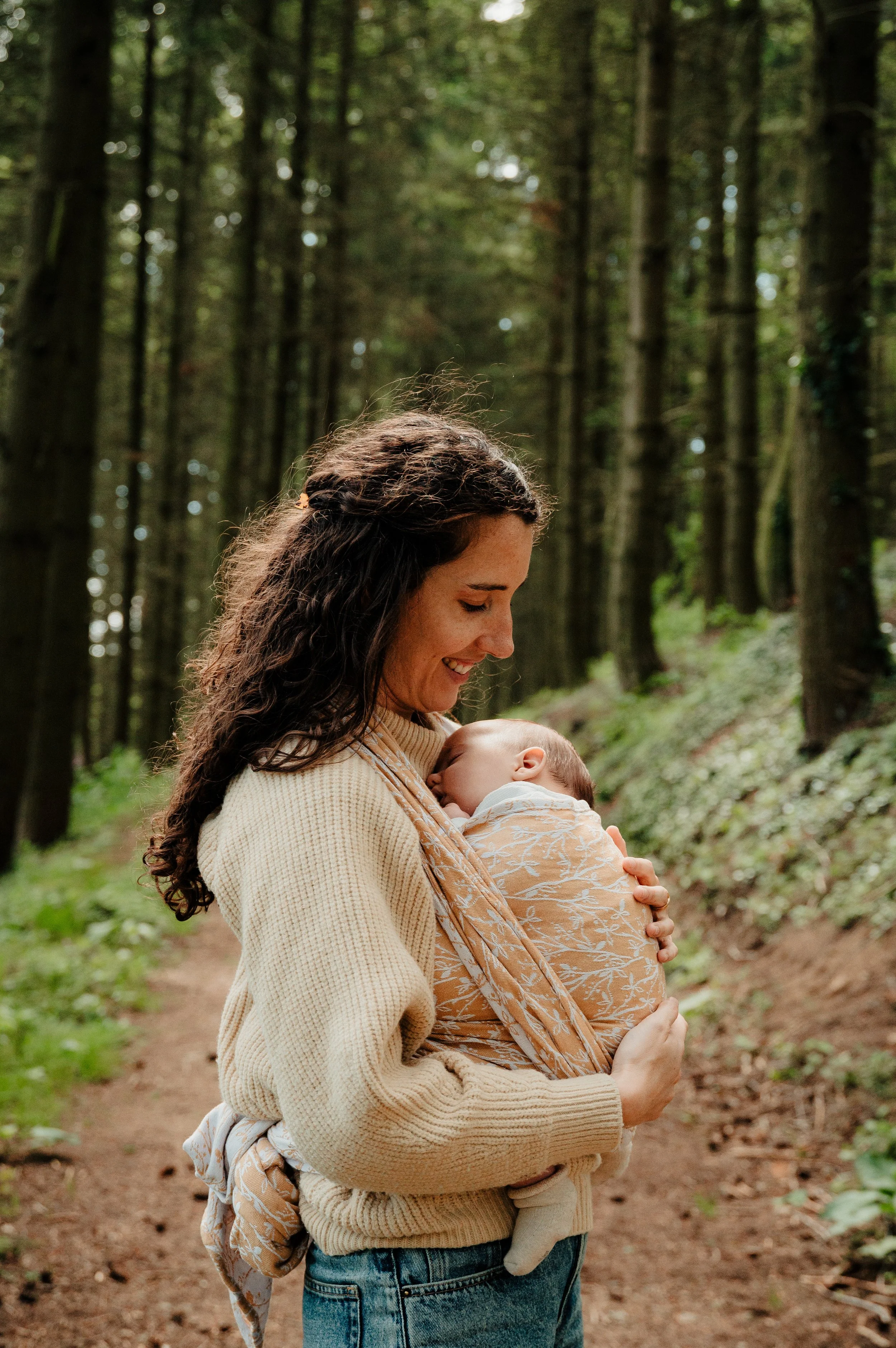 Une femme souriante dans une forêt, tenant un bébé contre elle, tous deux près d'un sentier rustique.
