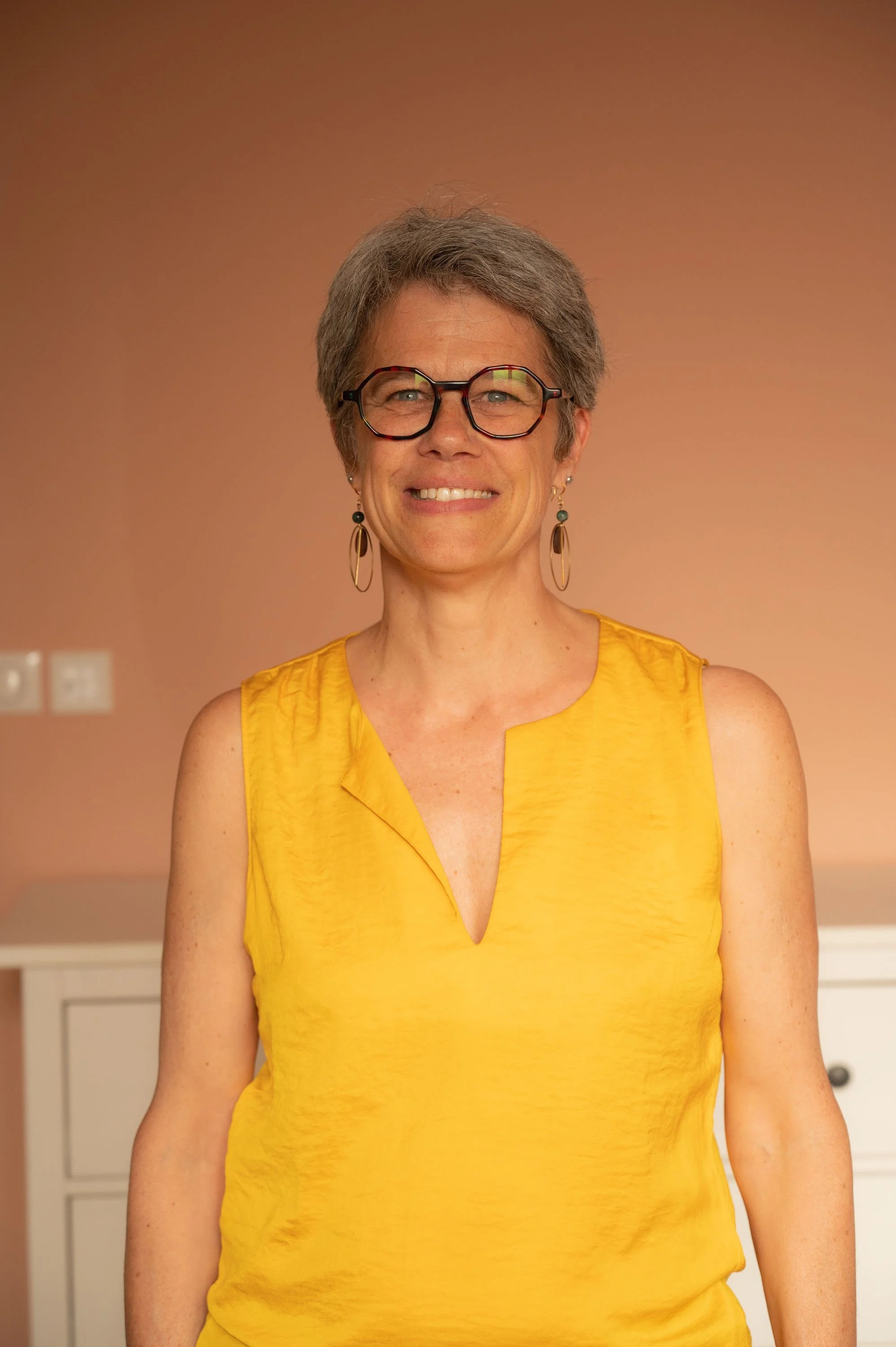 Une femme souriante avec des lunettes, portant une robe jaune, dans un intérieur avec un mur beige en arrière-plan.