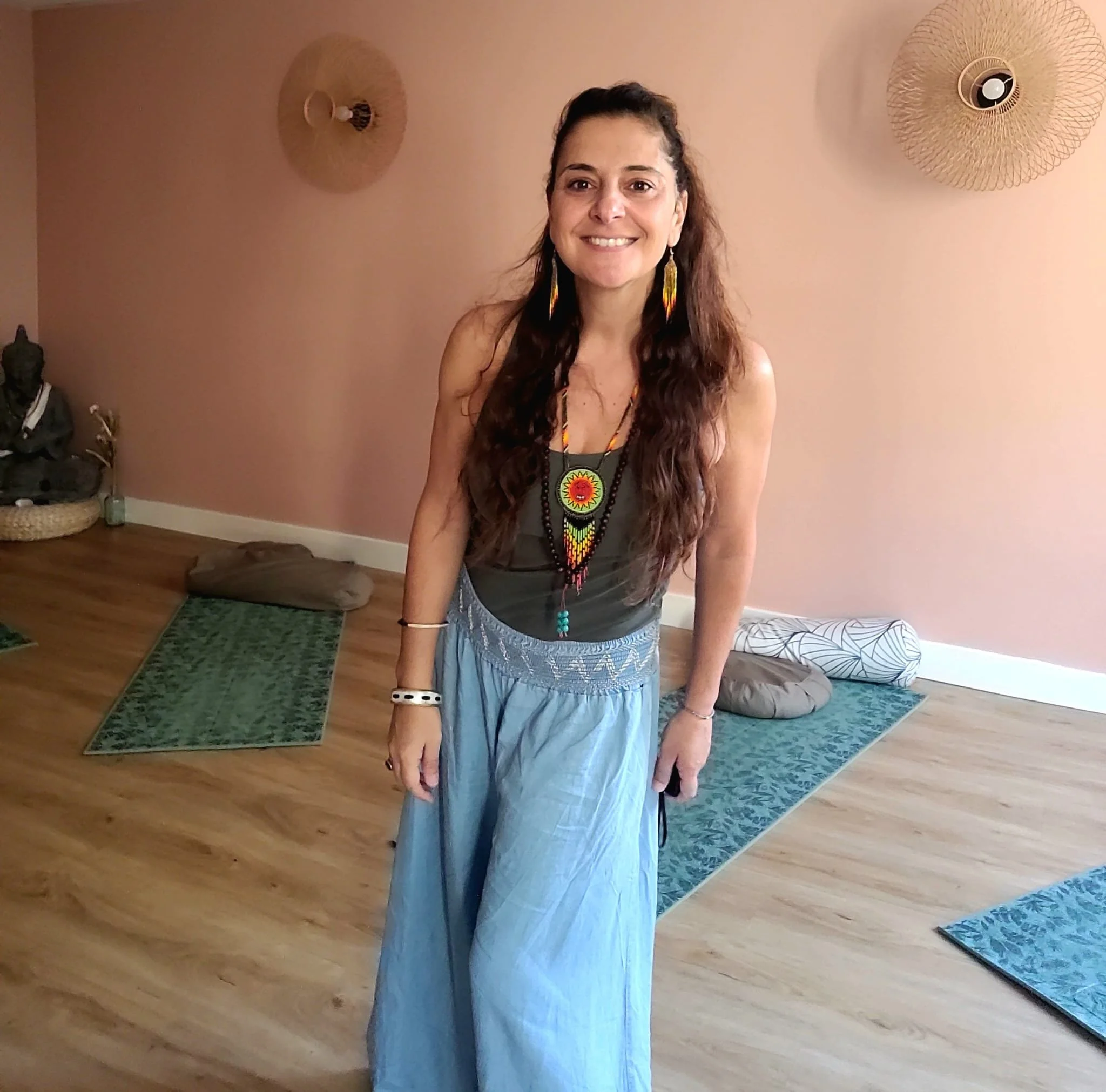 Une femme souriante portant des boucles d'oreilles longues et un collier avec un pendentif coloré, debout dans une pièce décorée avec des tapis en laine, une statue de Bouddha, des coussins et un mur peint en rose pâle.