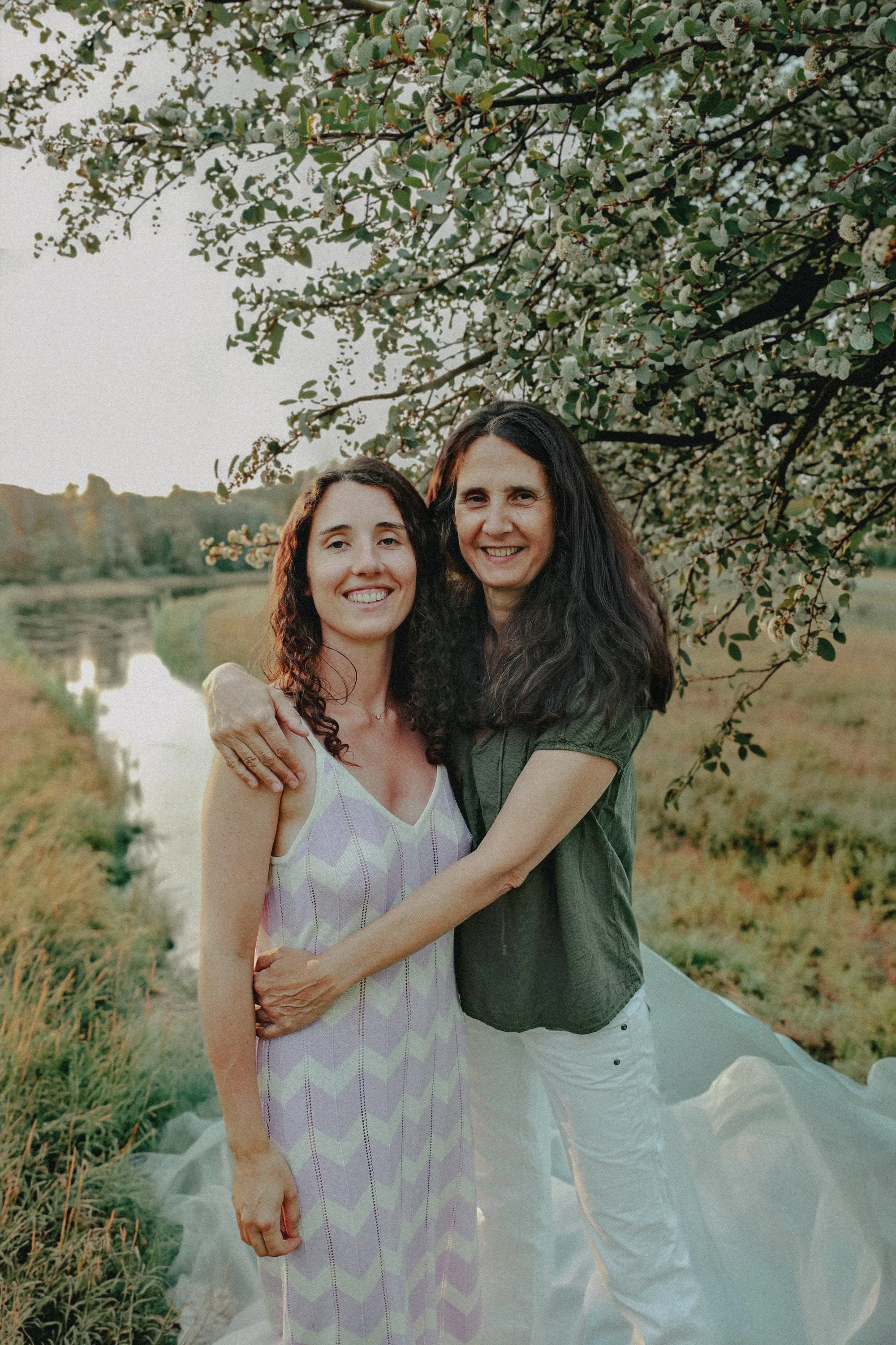 Deux femmes souriantes se tiennent près d'une rivière, entourées d'arbres en fleurs lors d'une journée ensoleillée.