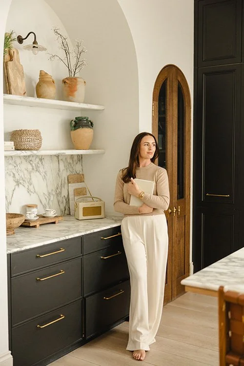 A woman standing in a modern kitchen, holding a notebook, with dark cabinets, open white shelves with decorative jars and plants, and a wooden arched door.