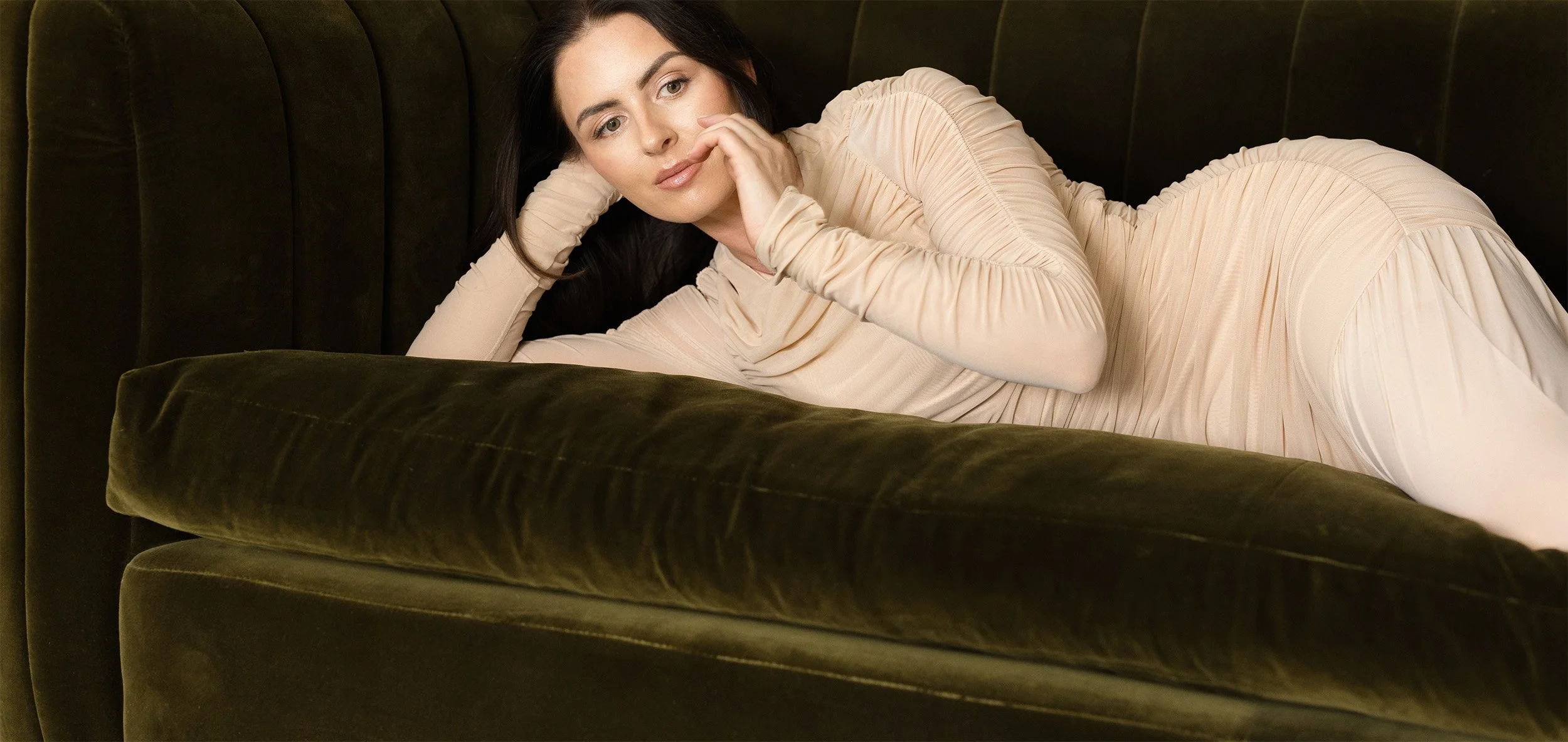 A woman with dark hair lying on a dark green velvet couch, wearing a light beige long-sleeve dress, with her head resting on her hand and looking into the distance.