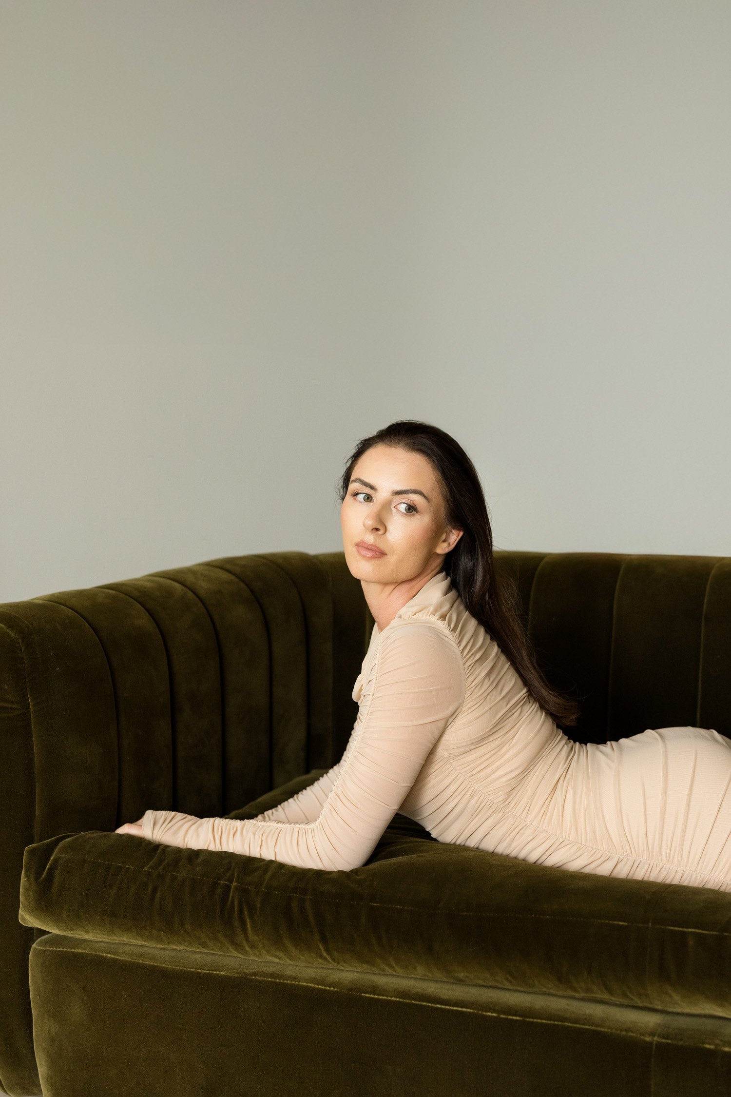 A woman with dark hair and fair skin, dressed in a beige long-sleeve dress, lying on her stomach on a vintage dark green velvet sofa against a plain light-colored wall.