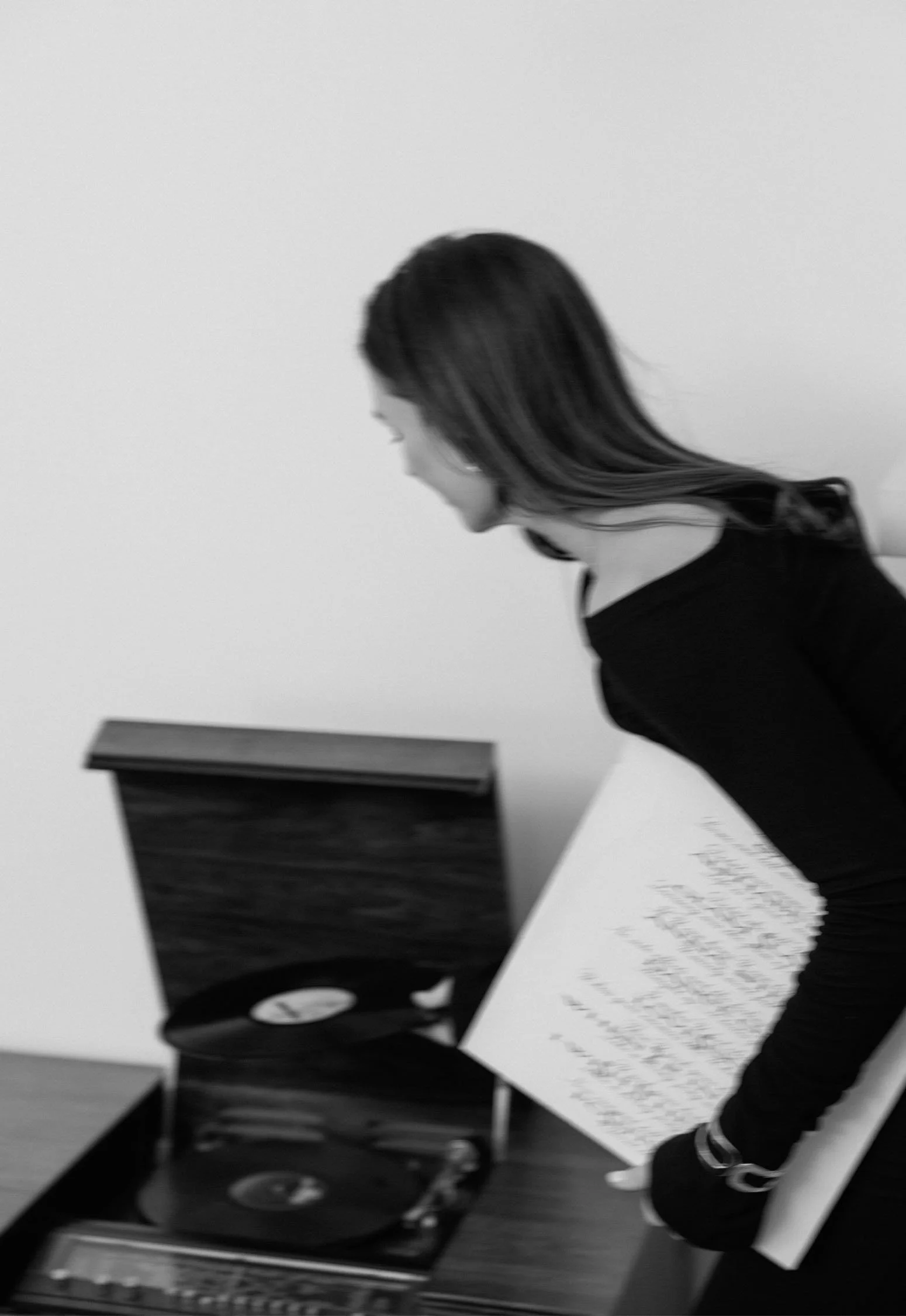 Black and white photo of a woman with long hair and glasses, standing beside a turntable and holding papers, looking down at the turntable.