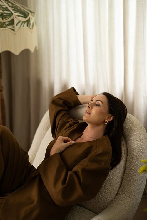 A woman relaxing on a beige armchair with her eyes closed, wearing a brown outfit, in front of a window with sheer curtains.