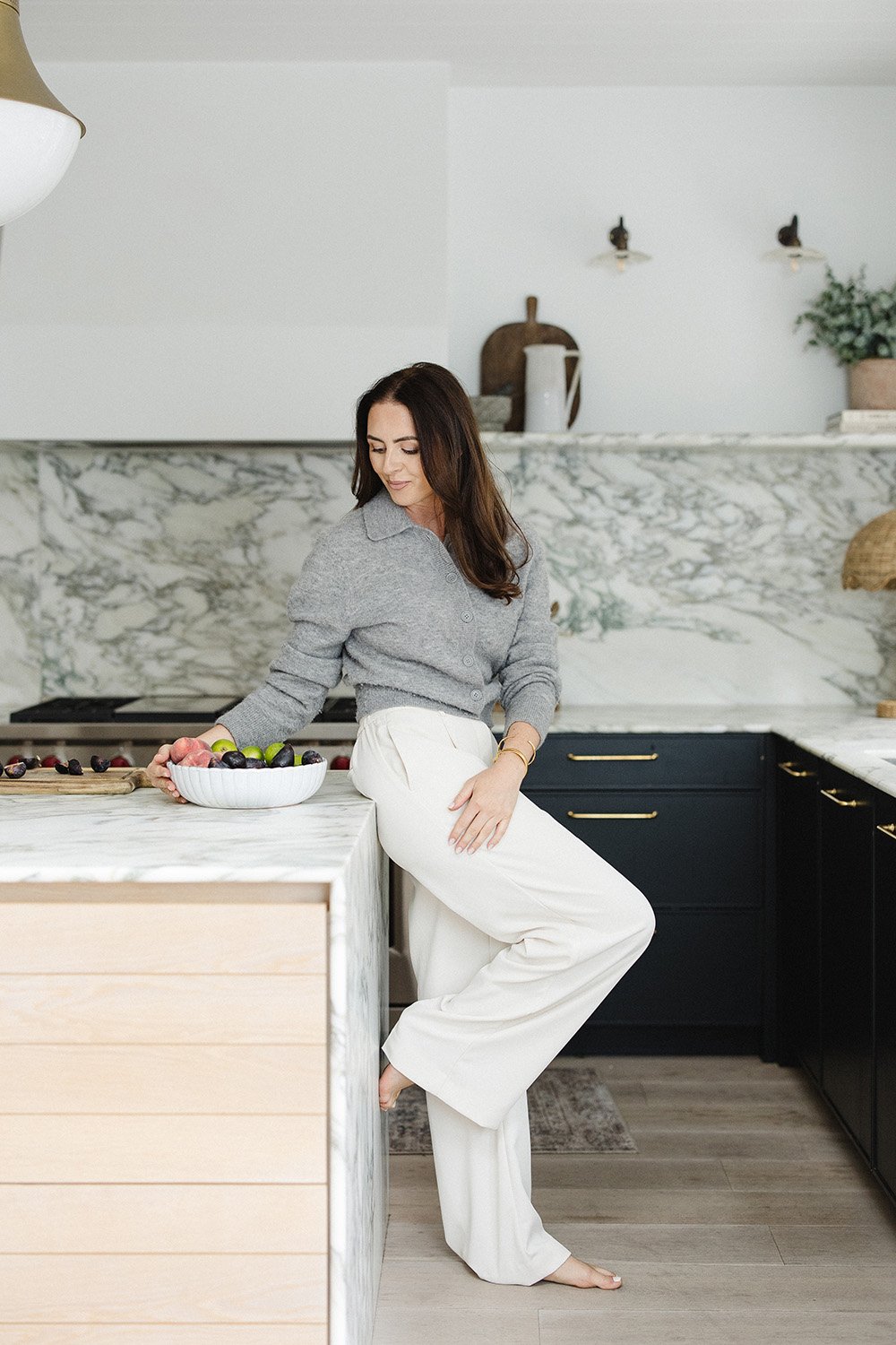 A woman in a gray sweater and white wide-leg pants sitting on a marble kitchen island, holding a bowl of assorted berries and fruits, in a modern kitchen with marble countertops and backsplash.