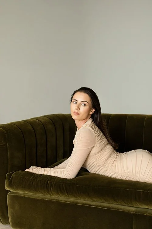 A woman in a beige dress is lying on a dark green velvet couch against a plain light gray wall.