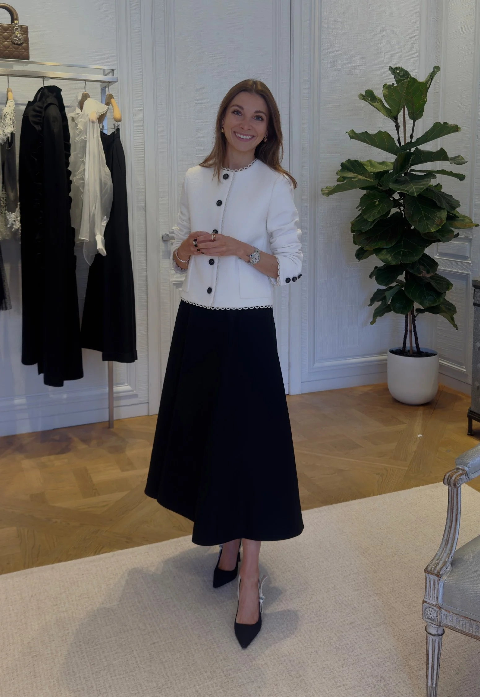 A woman in a white jacket and black skirt posing indoors, with clothing on hangers and a large potted plant in the background.