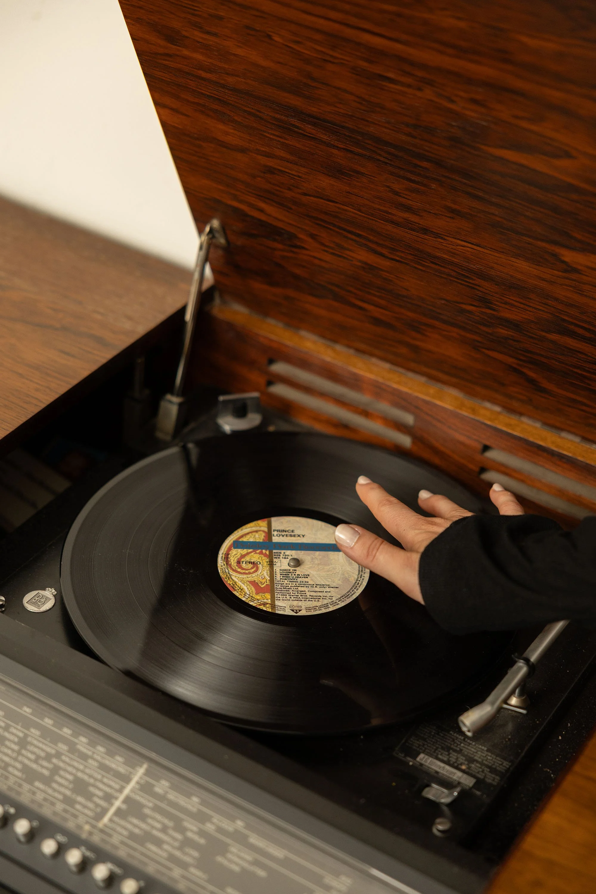A person with a black sleeve touching a spinning vinyl record on a turntable, with a wood-paneled lid lifted open.